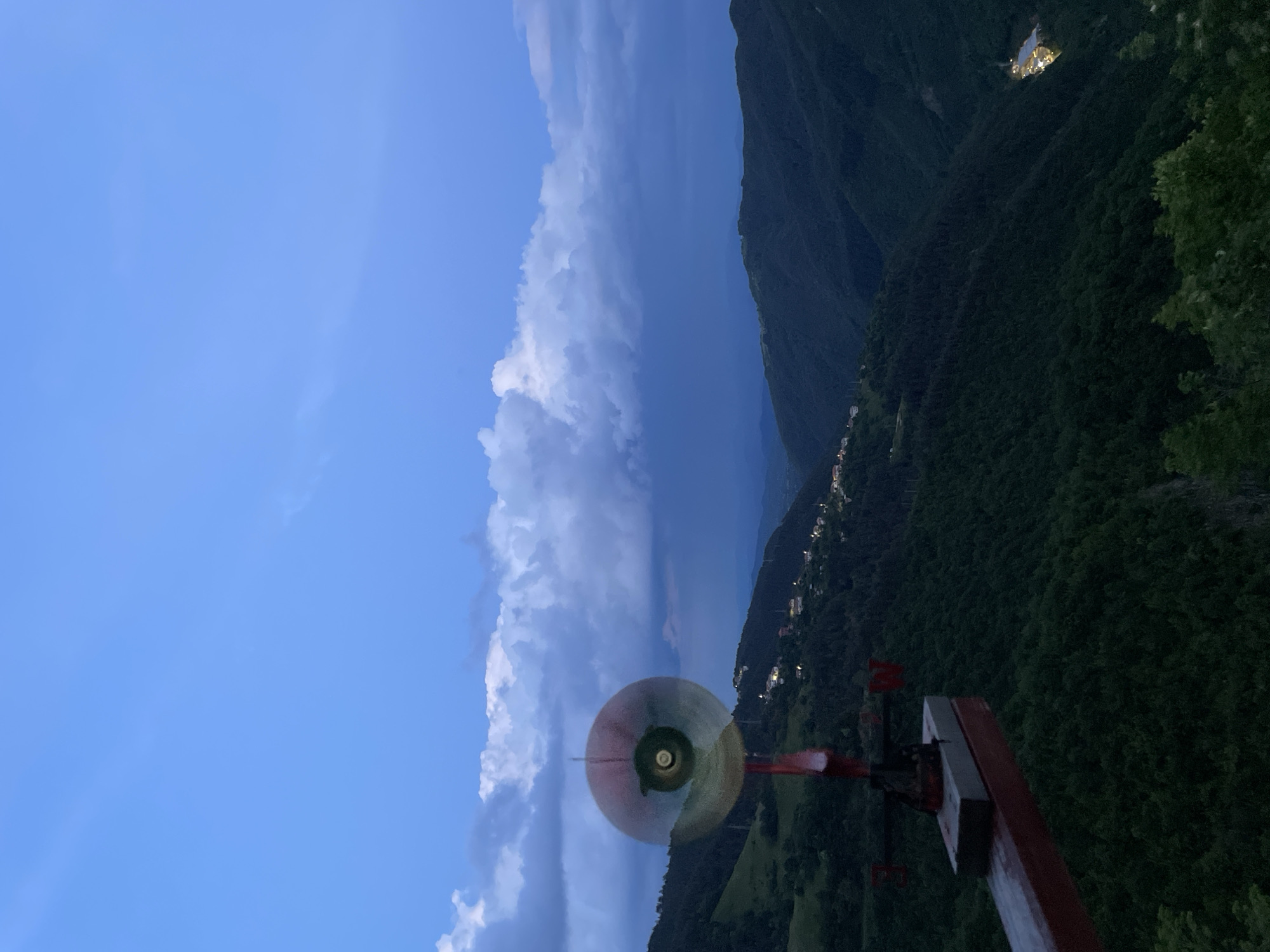 The image depicts a landscape featuring mountains, a sky with clouds, and an object in the foreground resembling a weather vane or a specialized antenna mounted on a wooden structure. The sky shows a gradient from light blue to darker, stormier clouds. Mountains are visible in the background, covered with vegetation and some evidence of settlements or buildings. The foreground object consists of a circular, partially reflective piece, mounted on a red and gray cross-shaped mechanism, itself atop a wooden base. The location may be Pechinska, Bulgaria, based on associated geographical and textual context.