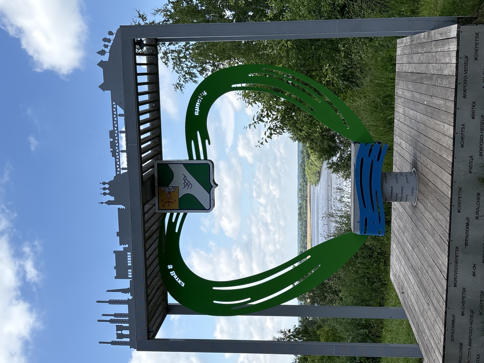 This is a photograph of a large, green, heart-shaped sculpture in the city of Kirovo-Chepetsk, Russia. The sculpture is made of metal and has cutouts within its shape. At the top of the sculpture is a metal silhouette of various buildings, including what appear to be industrial structures and a church. A coat of arms with green and white wavy lines and a yellow object is mounted within the heart shape. The sculpture is situated on a wooden platform, and a blue bench-like structure with wavy patterns is placed inside the lower part of the heart. The background shows a wide river with trees and vegetation on its banks, under a partly cloudy blue sky. The words "#УРАЛХИМИЯ" and "#КИРОВО-ЧЕПЕЦК" are visible on the base of the platform. The scene is outdoors, with lush greenery surrounding the sculpture.