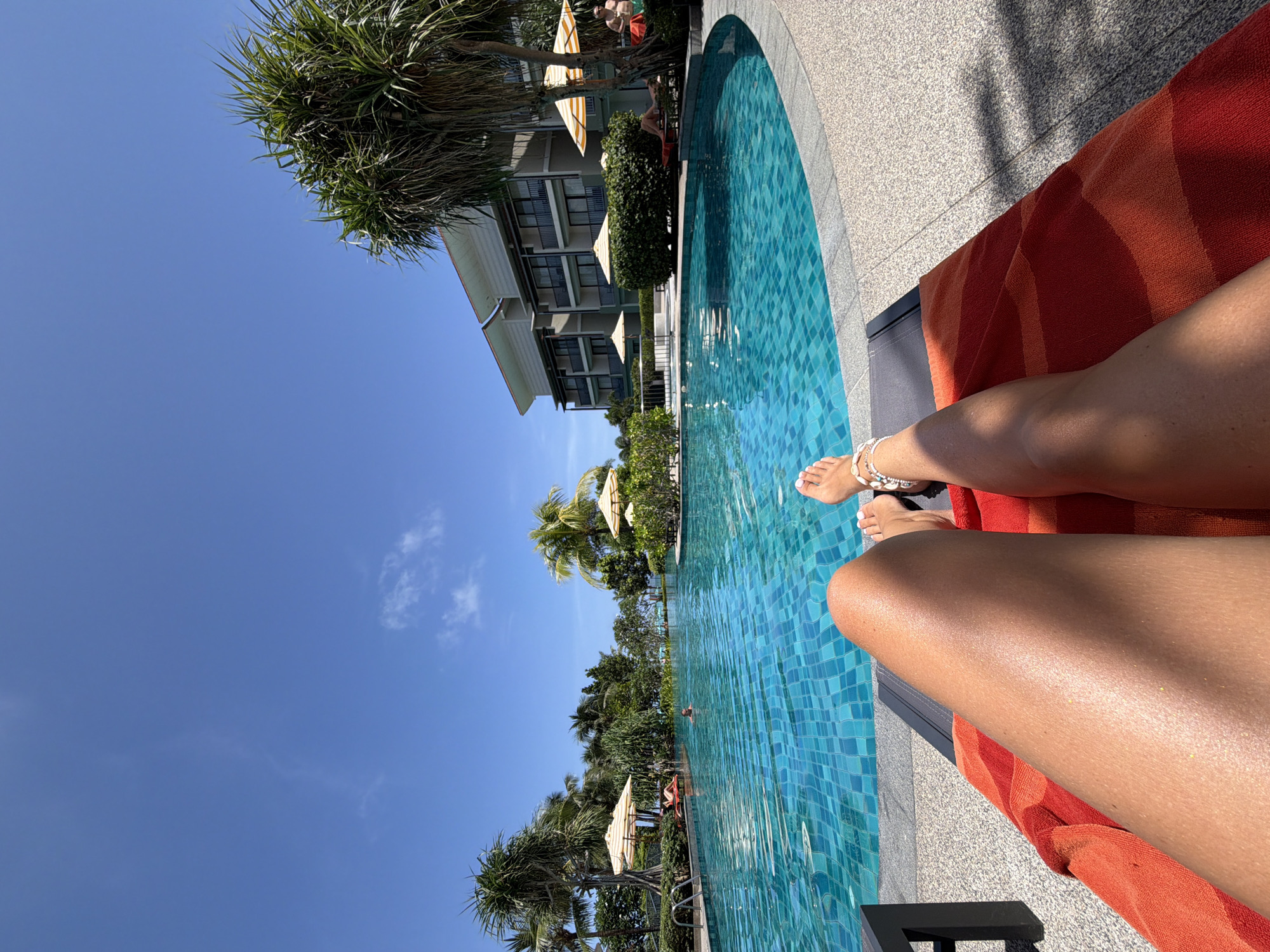 A first-person perspective shot captures a sun-drenched scene at a resort pool in Bang Muang, Thailand. The camera is positioned low, focusing on the legs of a person lounging on an orange towel atop a dark-colored lounge chair. The individual wears a white anklet with shell-like beads. To the right, another person's feet are visible, also adorned with a similar anklet.
The expansive, bright blue pool dominates the mid-ground, its surface shimmering under the sunlight and revealing a mosaic of blue tiles beneath. Several beach umbrellas with yellow and white stripes dot the poolside, providing shade. Lush green palm trees and other tropical foliage surround the pool, interspersed with a modern, multi-story hotel building in the background. A few other individuals are visible in the pool and relaxing on lounge chairs further away. The sky is a clear, vibrant blue with a few wispy clouds. The overall ambiance is one of relaxation and vacation.