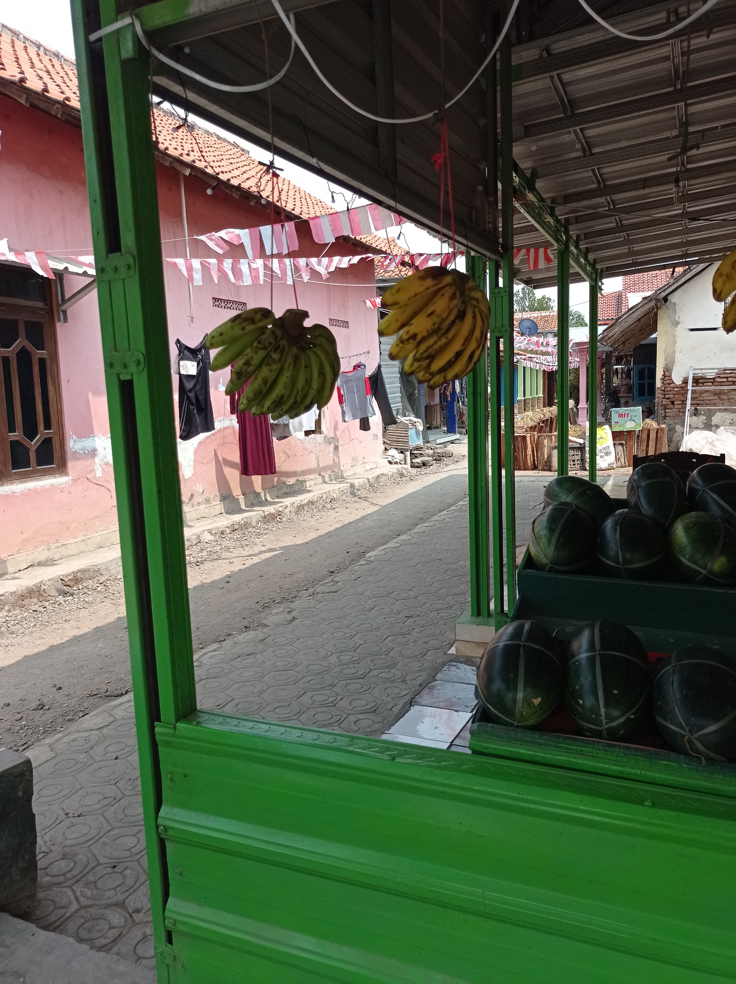 The image depicts a street-side fruit stand in Kramat, Indonesia. The stand is constructed with green metal framing and a corrugated metal roof. Several bunches of green and yellow bananas hang from the structure. Below, a display of watermelons, banded with string, is arranged in green crates. In the background, a pink building with a tiled roof is visible, along with laundry hanging to dry. The ground is paved with hexagonal tiles, and a dirt path runs alongside. A narrow street recedes into the distance, lined with other buildings and market stalls. The scene is illuminated by natural daylight, casting shadows on the ground. There are no people clearly visible in the foreground.