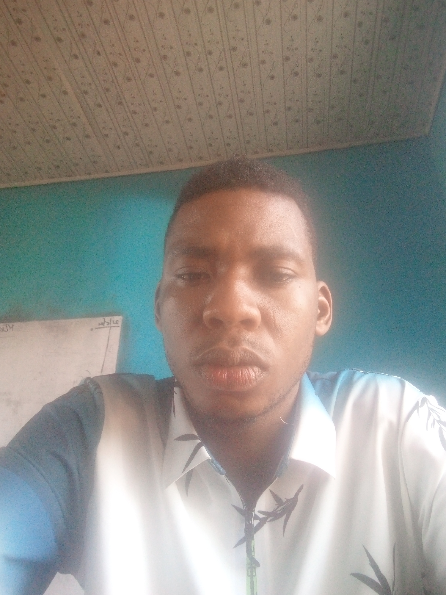 A medium-shot, face-on, selfie taken in an indoor setting. The subject is a dark-skinned man with short, dark hair. He is wearing a white collared shirt with a blue and black leaf pattern and blue sleeves. Behind the man is a blue wall. To the left of the man is a whiteboard with writing on it. Above the man is a white ceiling with a patterned design. Based on the prompt, this scene is located in Ikorodu, Nigeria.