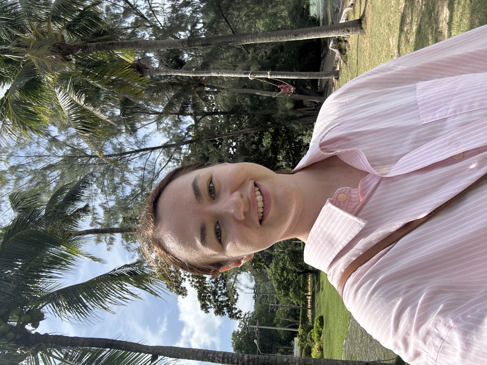 A medium-shot selfie shows a young Caucasian woman with shoulder-length brown hair and fair skin, smiling with her teeth showing. She is wearing a pink and white striped button-down shirt and a brown strap is visible across her shoulder. The background is a tropical landscape with tall palm trees, green foliage, a blue sky with scattered clouds, and a body of water. The location appears to be a park or beach area in Pa Khlok, Thailand, indicated by a sign in the background that partially reads "PALM COVE RESORT". The woman is positioned in the foreground, slightly to the left of center, with the lush vegetation filling the rest of the frame.