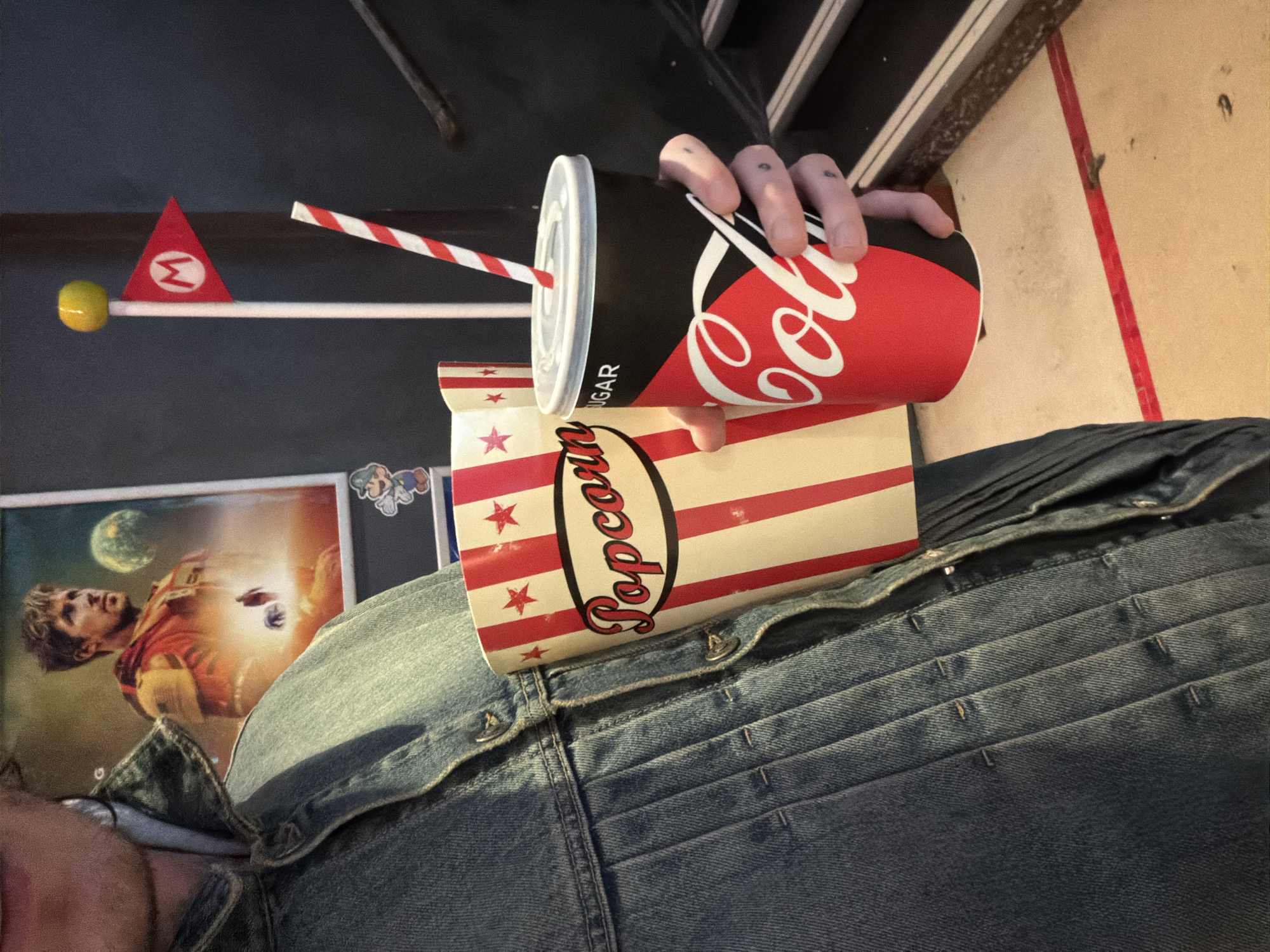A medium shot depicts a person seated, wearing a denim jacket, with a Coca-Cola cup in their right hand and a popcorn bucket resting on their lap. The cup has a red and white striped straw, and the popcorn bucket is adorned with red stripes and stars. In the background, a poster featuring a man in an orange and red outfit is visible, along with a smaller Mario-themed decoration. The setting appears to be indoors, possibly in a room with dark walls and light-colored flooring. It is not possible to identify the location as London from the given image.