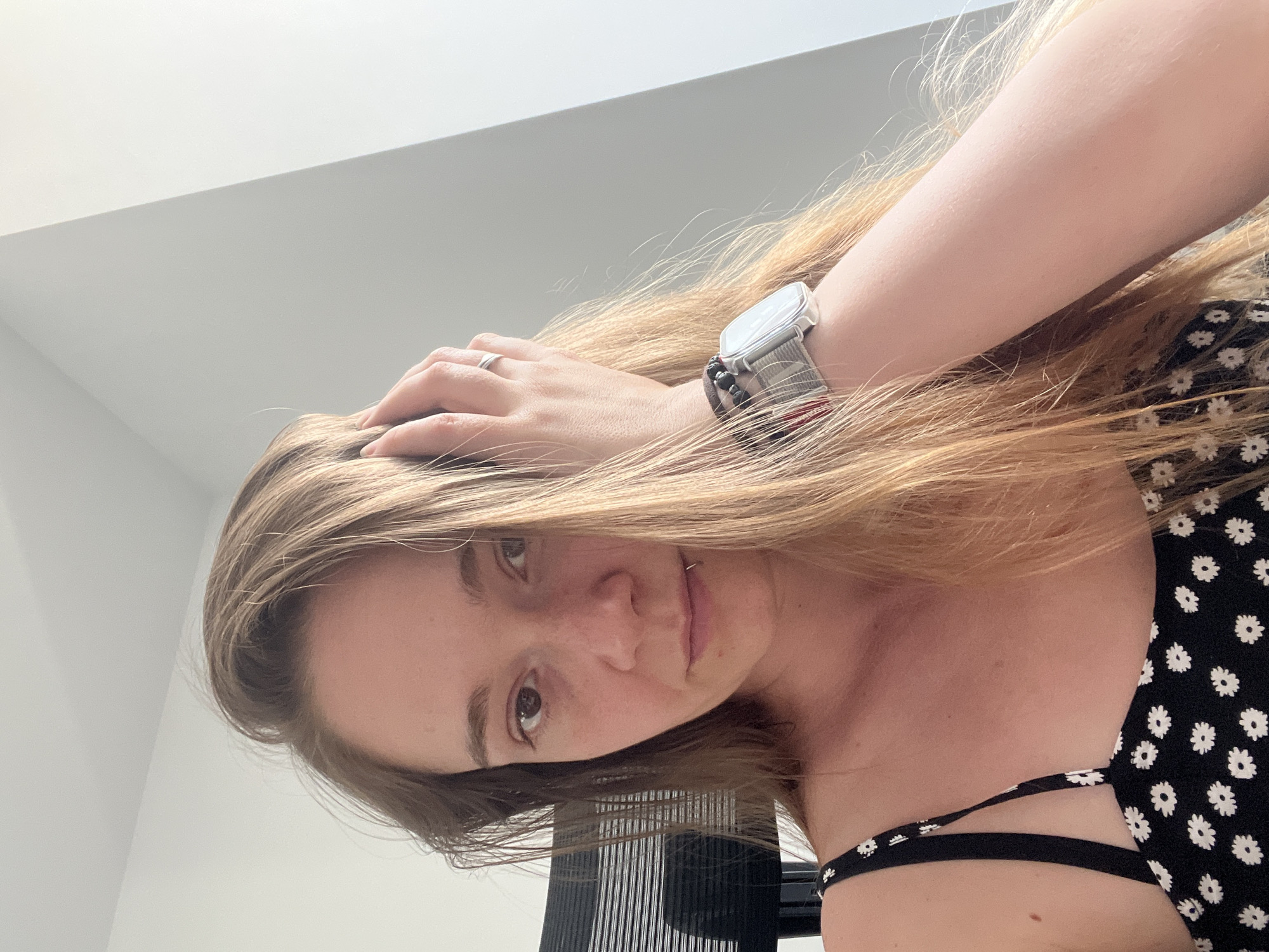 A woman with long, blonde hair is seated in an office chair, with her right hand raised to her head. She is wearing a black dress with a white floral pattern and a watch on her left wrist. The background consists of a white wall with a slanted architectural feature, and a black office chair is visible to her left. There are no discernible elements in the image that suggest a specific location in Plovdiv, Bulgaria.