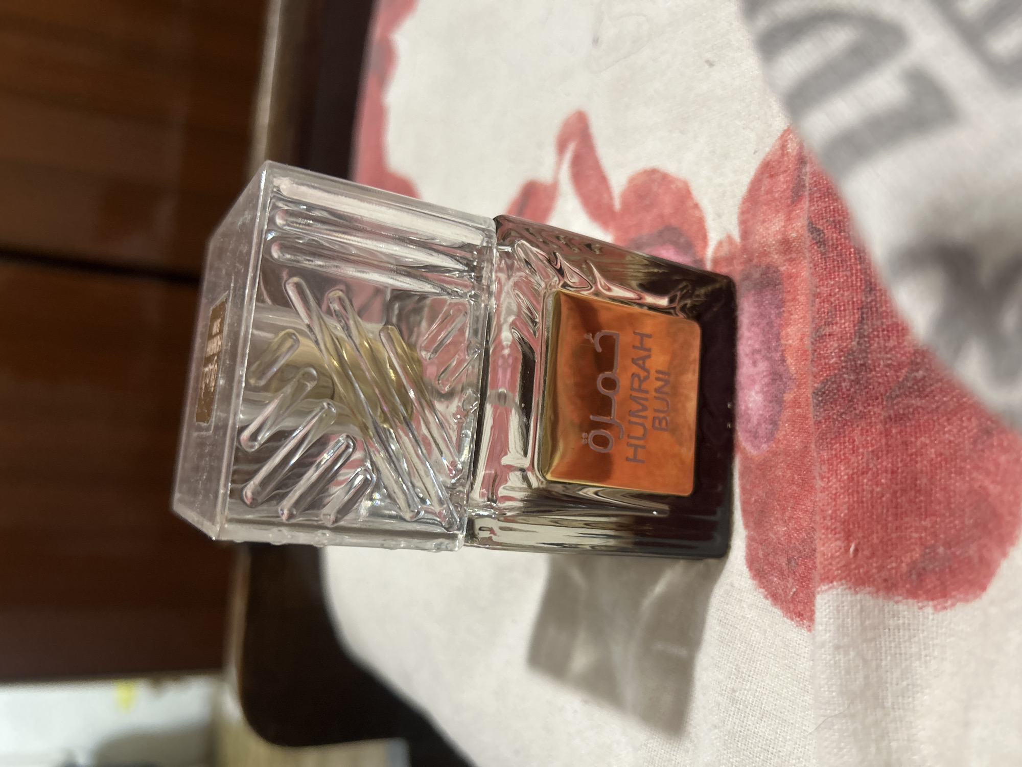 A clear glass perfume bottle with a textured cap sits on a white cloth with red floral patterns. The bottle has a golden label with Arabic script and the words "HUMRAH BUNI" printed in orange. The background is a blurred wooden surface. The image is taken from a slightly elevated angle. The location is not identifiable as Perushtitsa, Bulgaria.