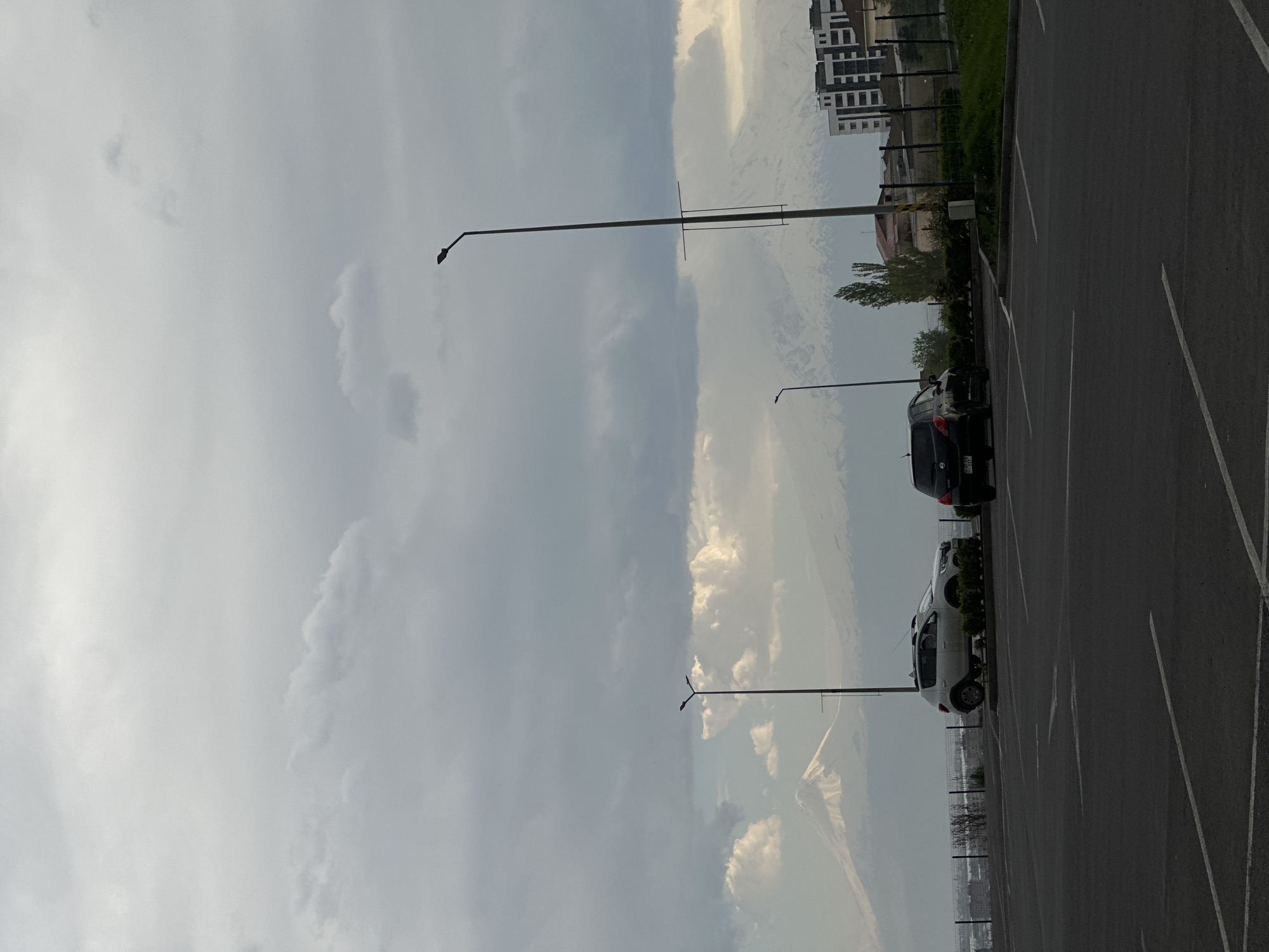 The image depicts a parking lot with parked cars under a cloudy sky. The location appears to be in an urban area with buildings visible in the background. There are lampposts interspersed in the scene. The parking lot is marked with white lines indicating parking spaces. There are multiple cars. The scene is outdoors during daylight.