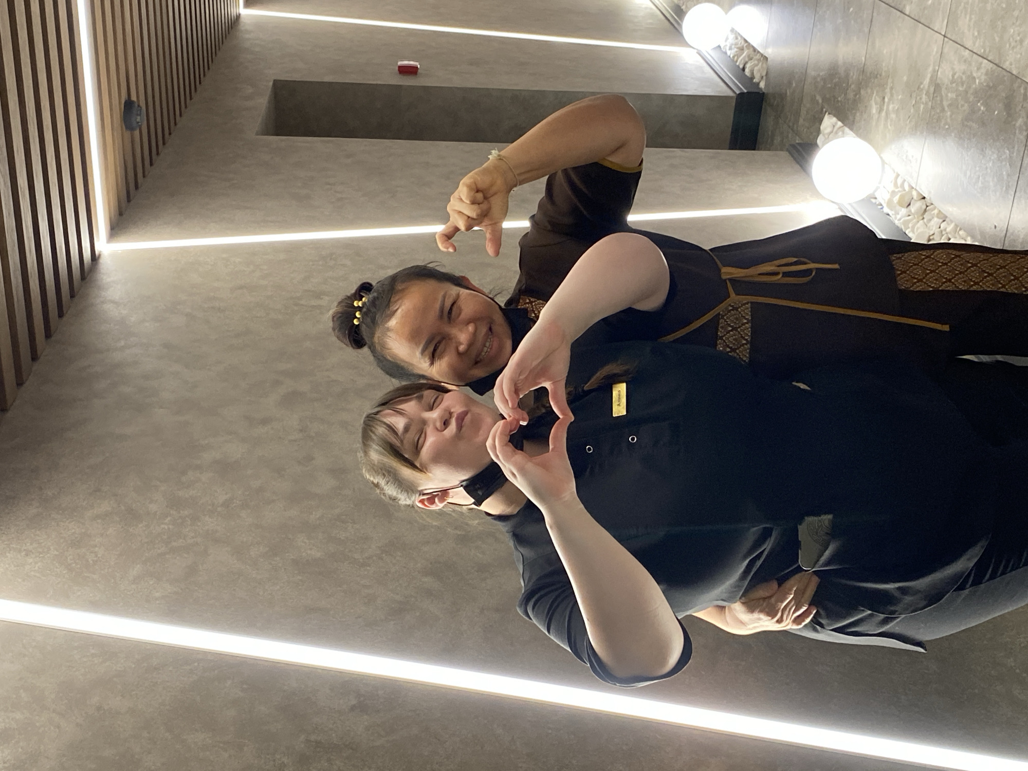 Two women are posing for a photograph in a modern indoor setting. The image is rotated, showing the subjects and background from an angled perspective.
The woman on the left (when the image is viewed upright) has dark hair styled in a bun and is wearing a dark brown uniform with gold-colored trim and an ornate patterned fabric around the waist and sleeves, reminiscent of traditional Thai attire. She is smiling broadly and forming a "finger heart" gesture with her hands.
The woman on the right has light brown hair with bangs and is wearing a black collared uniform. A yellow nametag pinned to her uniform reads "Alina". She has a black face mask pulled down to her chin and is creating a heart shape with both hands held in front of her chest. Her eyes are closed, and she has a gentle smile.
The background features contemporary interior design elements. Walls are a light, textured grey. Bright, linear LED lights are embedded along the edges of the walls and ceiling, providing illumination. On one side, there are wooden slatted panels, and on the opposite side, a decorative ledge filled with white stones and featuring two spherical white lights. A small red rectangular object is visible on a wall in the background.
Both women appear happy and friendly, making gestures of affection. The uniforms and modern, clean aesthetic suggest a professional environment, possibly a spa, salon, or wellness center.