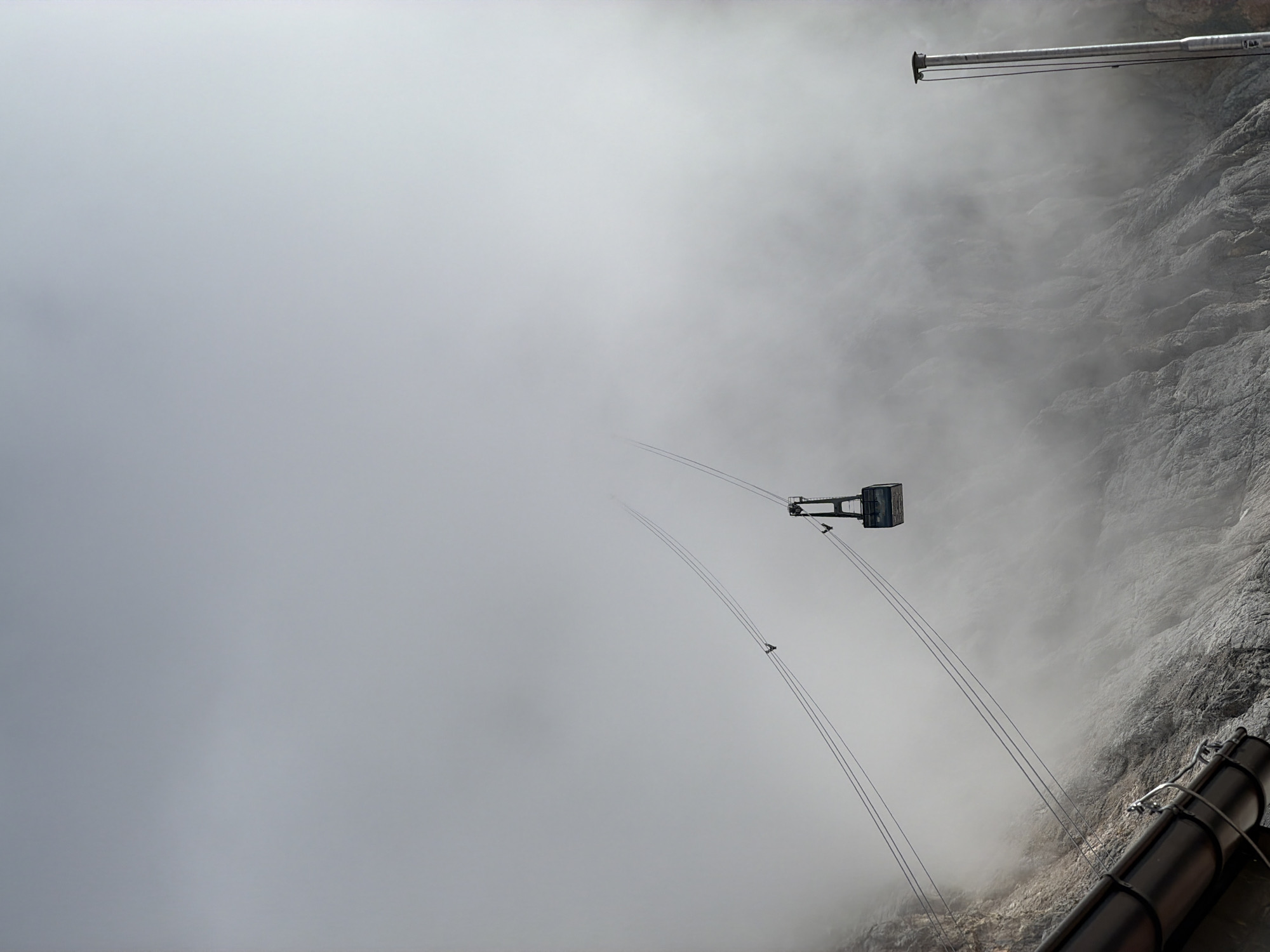 This is a vertical, medium-shot, eye-level photograph of a cable car ascending a mountain shrouded in fog, likely the Zugspitze in Garmisch-Partenkirchen, Germany. In the foreground, a dark, metallic gutter-like structure runs horizontally along the bottom left of the frame. The mountain face, covered in rocky terrain and patches of fog, occupies the lower half of the image. Two sets of parallel cables descend diagonally from the upper right to the lower left of the frame. A dark blue and grey cable car, suspended from the cables, is visible in the mid-ground, ascending towards the top of the frame. Two small, dark figures, presumably birds or people, are perched on the cables in the mid-ground. A tall, thin metallic pole with a cap extends vertically from the bottom right corner of the frame. The sky is overcast and filled with dense white fog, obscuring the upper portions of the mountain and the sky.