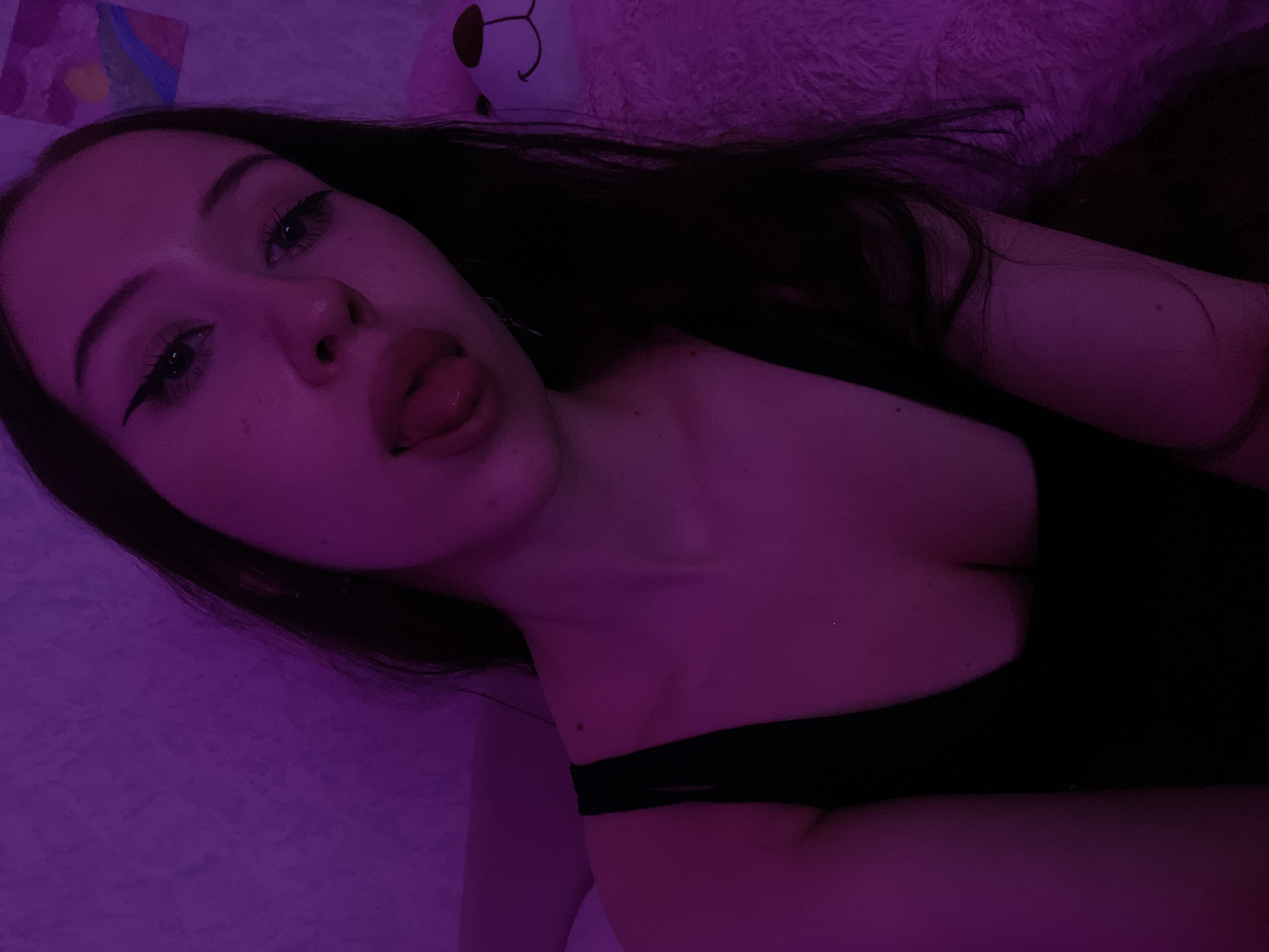 The image shows a close-up of a young woman with dark hair, wearing a black top. She is sticking her tongue out, and her eyes are lined with winged eyeliner. The scene is illuminated with purple light, which casts a shadow on the wall behind her. To the right of the woman is a large, fluffy, white teddy bear. The background wall has a textured surface. A small, colorful painting is visible on the wall. The image appears to be taken indoors, in a bedroom or personal space. There is no information available to determine the location within Votkinsk, Russia.