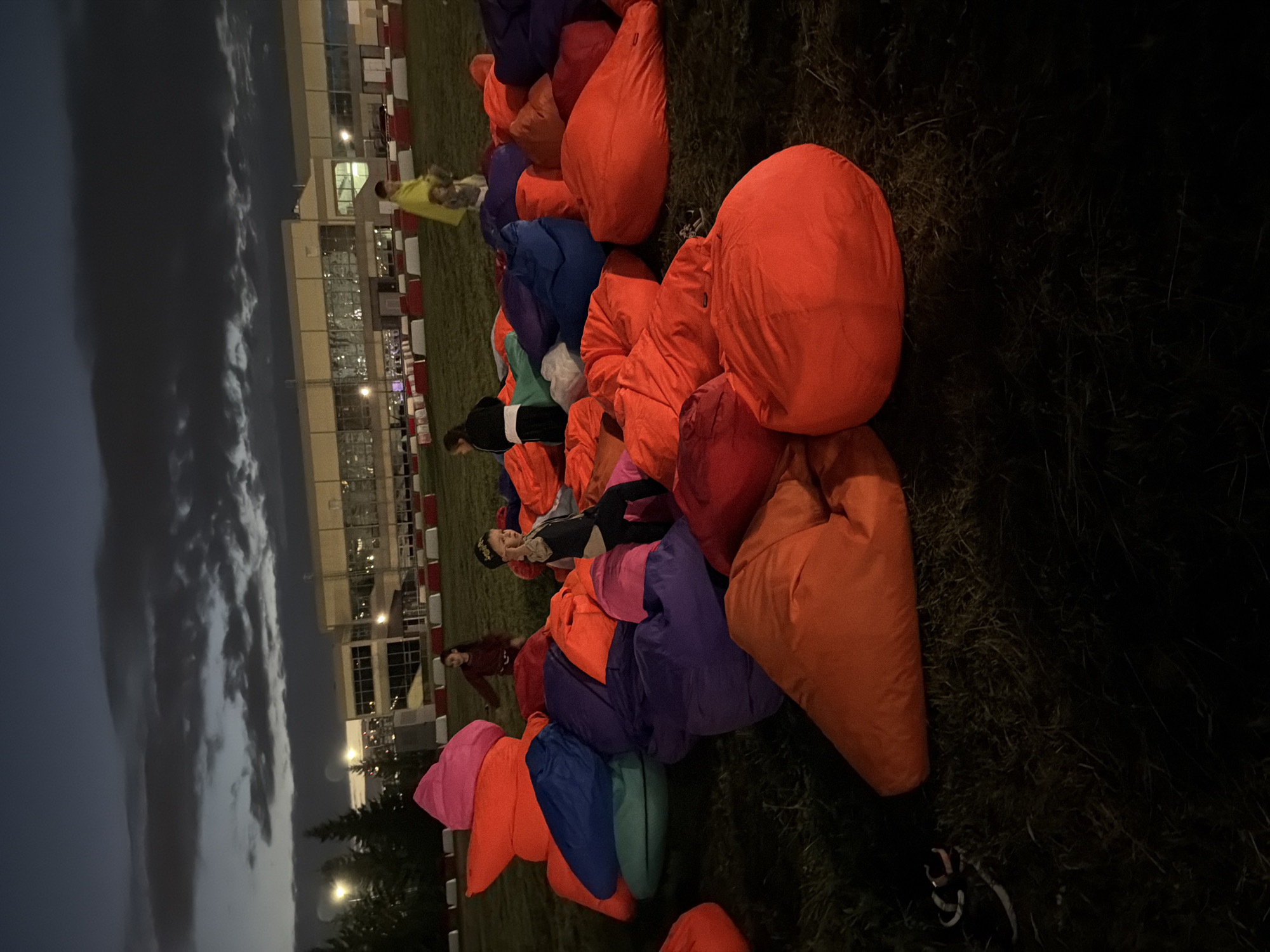 A group of children are gathered in a grassy area in front of a long, low building with many windows. It is dusk, and the sky is cloudy. The children are sitting on or playing with a large collection of brightly colored beanbag chairs. Some of the beanbag chairs are stacked high. In the background, there are some trees and streetlights. The building in the background appears to be an industrial or commercial building. There are also red and white barriers in front of the building, which could indicate a race track or other event space.