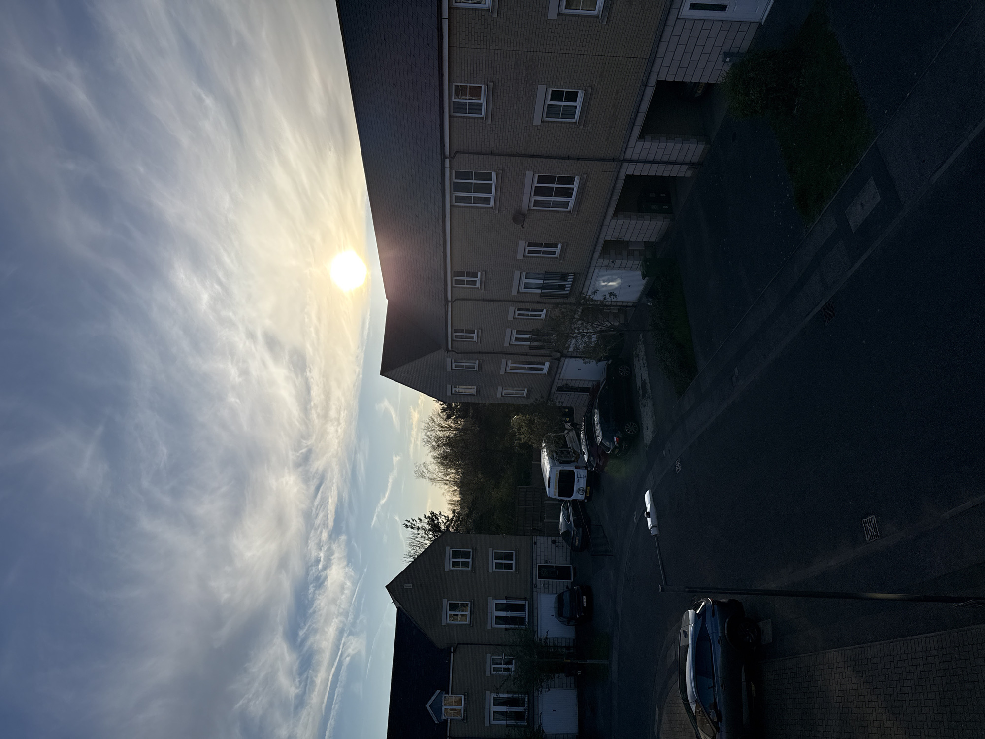 The image depicts a residential area in Great Yarmouth, United Kingdom. The view is elevated, possibly from an upper floor of a building. The scene includes several multi-story apartment buildings with light-colored brick facades and dark roofs. Numerous windows are visible on each building. A street runs between the buildings, with parked cars and at least one van visible. Trees are interspersed throughout the scene, adding some greenery. The sky is partly cloudy, with the sun visible through the clouds. The lighting suggests it might be late afternoon or early evening. The overall impression is a quiet, suburban neighborhood.