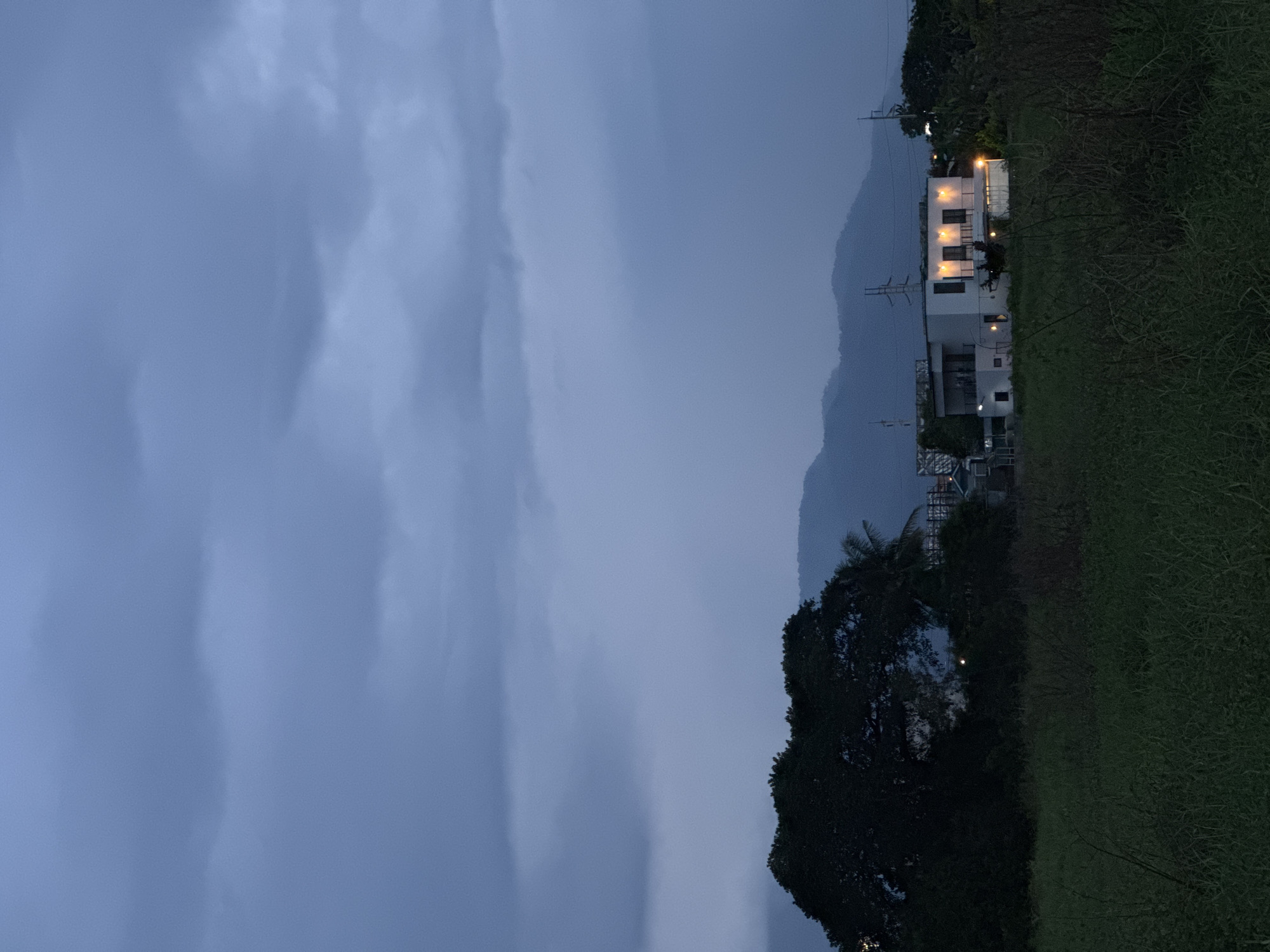 The image depicts a landscape in บ้านกาญจน์กนก 23, Thailand, during a cloudy evening. A multi-story white building with illuminated windows stands on the right, partially obscured by trees. Mountains are visible behind the building, fading into the cloudy sky. The foreground consists of tall green grass and dense foliage. The overall lighting is dim, emphasizing the darkness of the trees and the cloud cover.