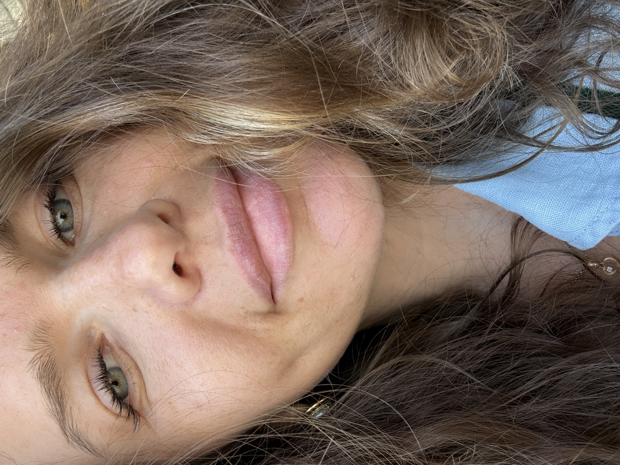 The image is a close-up, overhead shot of a woman's face, with focus on her eye, nose, and mouth. Her hair, a mix of brown and blonde, is messy and frames her face. She appears to be outdoors, with natural lighting illuminating her features. There is no discernible location in the city of Dubai, United Arab Emirates, within the image. The woman is wearing a light blue collared shirt and a small, heart-shaped pendant necklace.