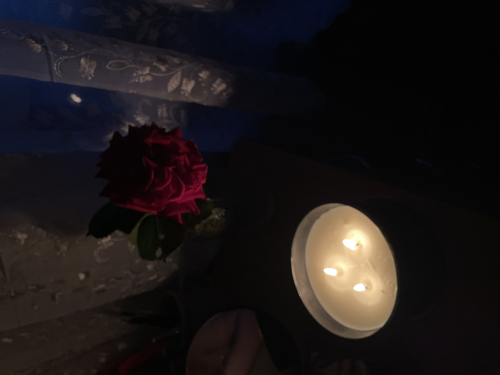 The image presents a dimly lit, intimate indoor scene featuring a vibrant red rose and a burning three-wick candle. The setting is dark, suggesting nighttime or a deeply shadowed room, with light primarily emanating from the candle flames, casting a warm, soft glow.
In the upper-left portion of the frame, a segment of a patterned, translucent white fabric, possibly a curtain or tablecloth with embroidered details, is visible. It reflects some blue light, contrasting with the warm candle glow. Below this fabric, a single red rose with dark green leaves stands out, its petals richly colored against the dark background.
To the right, a white three-wick candle, housed in a dark container, burns brightly. The flames are clearly visible, illuminating the wax surface. The candle is positioned at an angle, suggesting it sits on a reflective or dark surface.
There are no people or explicit activities depicted. The arrangement of the rose and candle creates a serene, contemplative, or romantic atmosphere. No visible text is present in the image. While the provided context indicates Zlatograd, Bulgaria, there are no specific visual cues within the image to confirm this location.