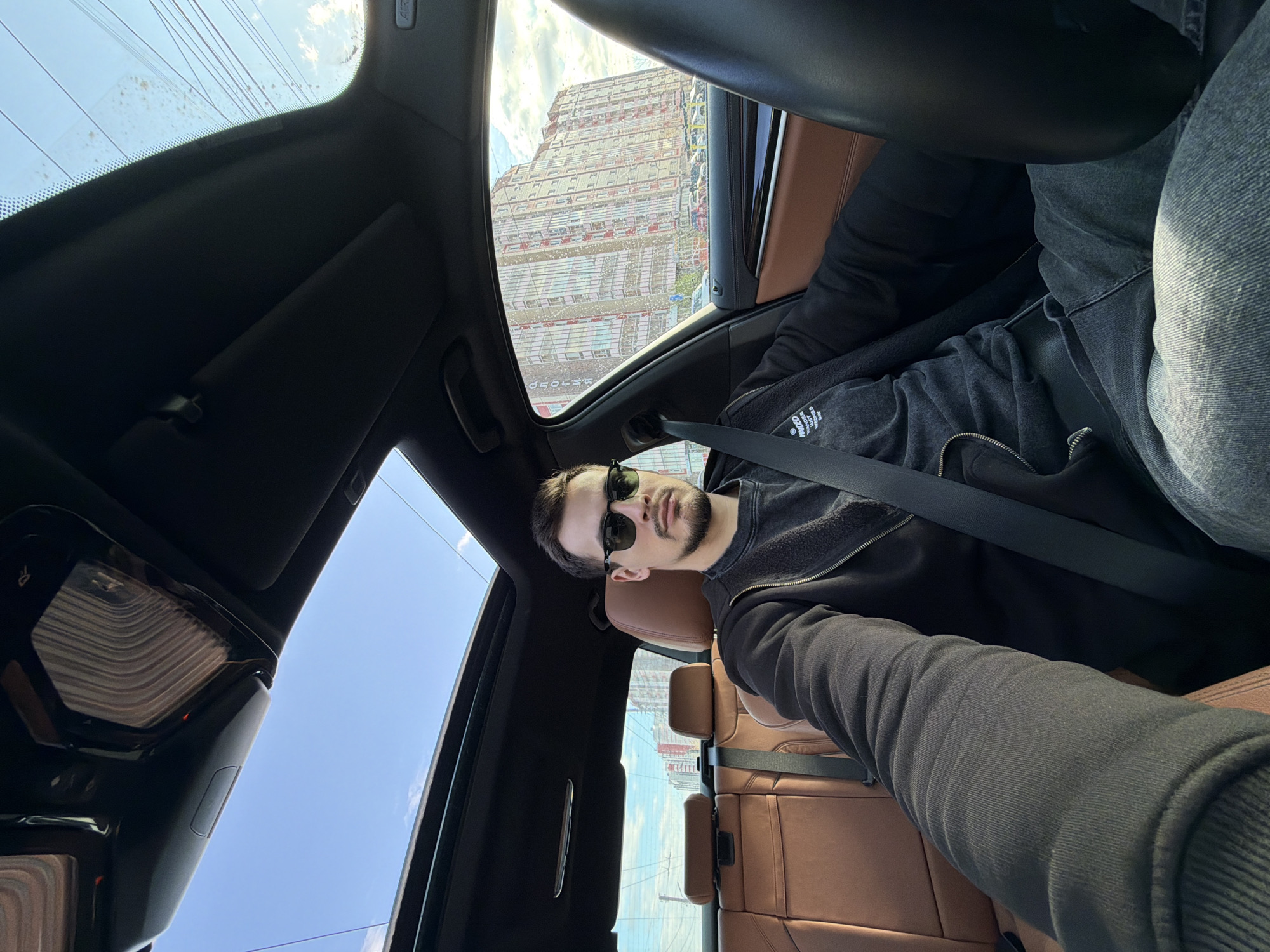 The image captures a man wearing sunglasses and a dark sweatshirt inside a car. He is wearing a seatbelt. The car's interior features brown leather seats and a sunroof. Outside the sunroof, a tall building under construction is visible. This building appears to be located in Krasnoyarsk, Russia.