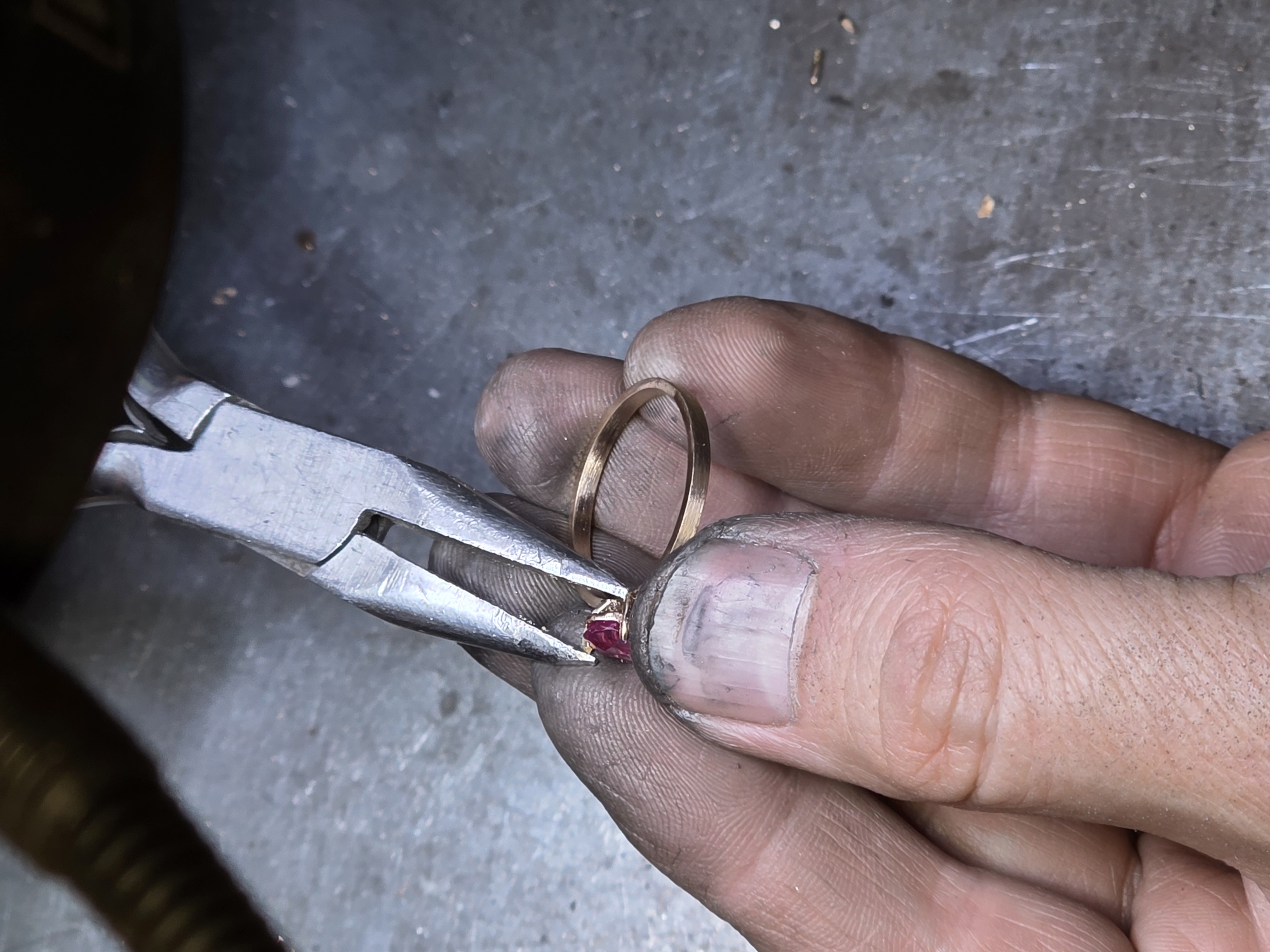 A close-up, overhead shot shows a pair of hands working on jewelry. A person's thumb and forefinger, coated in a gray dust, hold a ring and a marquise-shaped red gemstone. Pliers, made of silver-toned metal, grip the gemstone, appearing to set it into the ring. A second, plain gold ring rests on the person's ring finger. The background is a textured, dark gray surface, likely a workbench, with scattered metal shavings. This scene is not specifically located in Kazan, Russia, as there are no identifiable landmarks or contextual clues to indicate a geographical location.