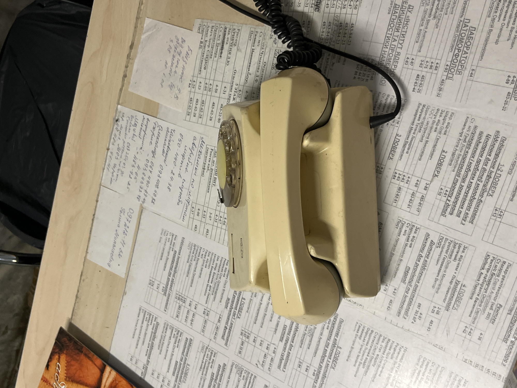 An old, cream-colored rotary dial telephone with a coiled cord lies on a surface covered with printed documents, possibly from a medical or research facility, suggesting a setting in Kyiv, Ukraine. The documents contain Cyrillic text and numerical data, some of which appear to be phone numbers or codes. The telephone's rotary dial is partially visible, and the handset rests on its base. The background is dark and out of focus, hinting at an indoor, possibly office or storage environment. One document has a handwritten note in Cyrillic that reads "097 812-71-36". Another document has "5 поверх" (5th floor) written on it. The overall impression is one of an older, perhaps disused, piece of technology placed on top of various informational papers.