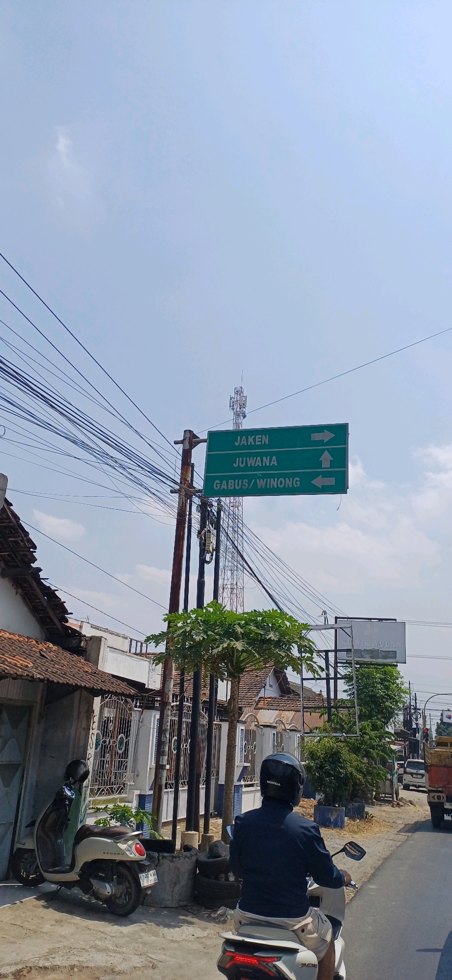 The image depicts a street scene in Jakenan, Indonesia. A green directional sign with white lettering indicates routes to "JAKEN" with an arrow pointing right, "JUWANA" with an arrow pointing up, and "GABUS/WINONG" with an arrow pointing left. The sign is mounted on a metal pole alongside a cellular tower. In the foreground, a person wearing a dark jacket and light-colored shorts rides a white scooter, identified by a "HONDA" logo on their helmet. To the left, a beige scooter is parked near a building with a red tile roof and a white fence. The environment includes a clear sky with sparse clouds, overhead power lines, trees, and other buildings in the background.