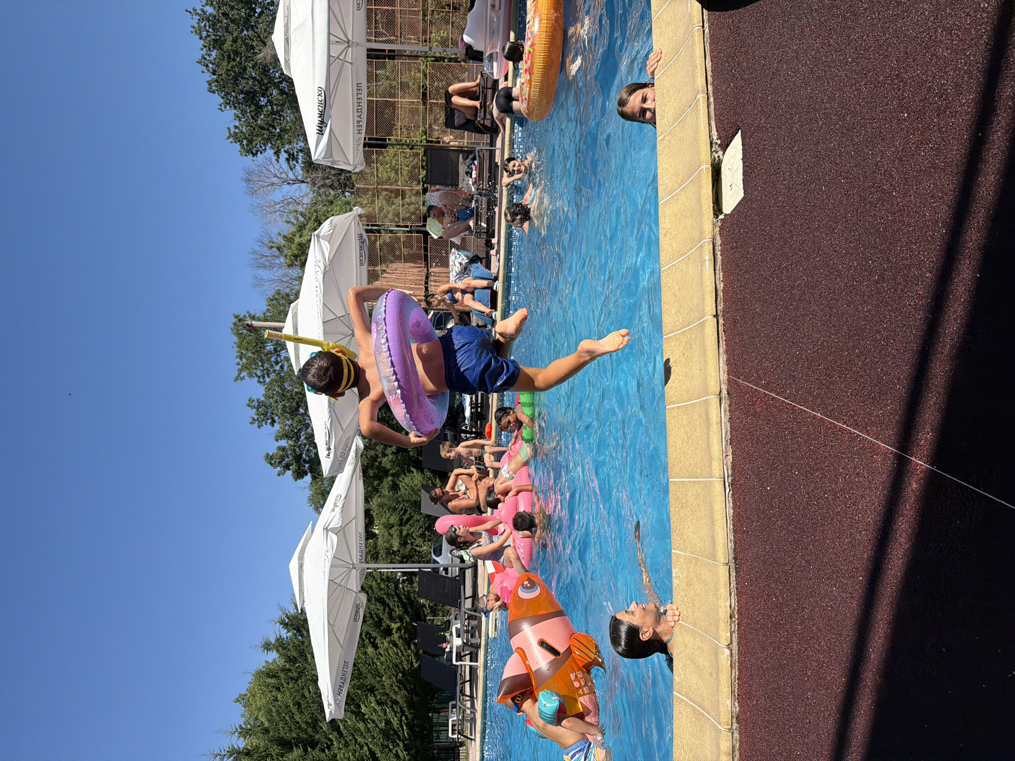The image depicts a busy summer day at an outdoor swimming pool facility, likely located in the city of Burgas, Bulgaria, given the context provided by the umbrellas featuring Cyrillic text. In the foreground, a young boy, wearing blue swim trunks and a yellow snorkel mask, is captured mid-air as he jumps into the pool, holding a purple inflatable ring. The pool is filled with people of various ages enjoying the water. Several individuals are seen swimming, while others are relaxing on inflatable toys, including a large pink flamingo float. In the background, lounge chairs and white umbrellas provide shade, suggesting a well-equipped recreational area. The overall atmosphere is one of leisure and fun on a bright, sunny day.