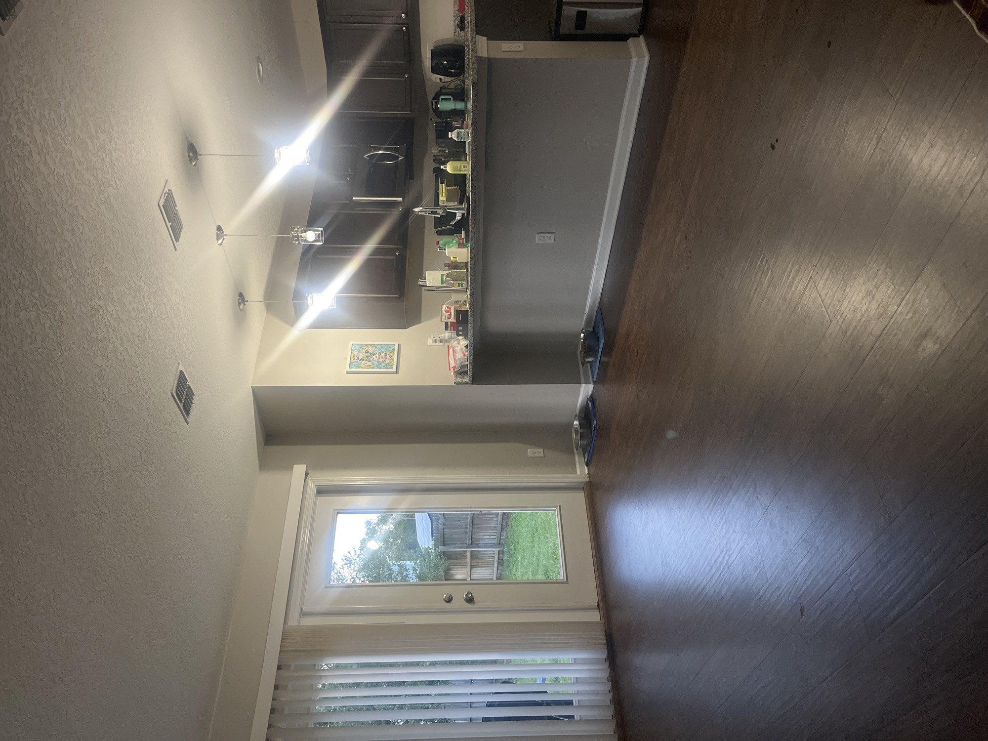 The image depicts the interior of a residential living area adjacent to a kitchen, with a view of a backyard through a glass door. The flooring is dark hardwood. In the foreground, the floor shows some smudges and reflections from overhead lighting. To the left, a glass door leads to a backyard with a wooden fence and green grass, partially obscured by vertical blinds. In the middle ground, a portion of the kitchen counter is visible, with dark cabinets above it. The counter is stocked with various items including bottles, containers, and kitchen appliances like a coffee maker and a black air fryer. There are three pendant lights hanging from the ceiling above the kitchen counter. On the floor near the kitchen counter, two pet bowls are placed on blue mats. To the right of the kitchen counter area, a silver trash can is visible. The walls are painted a neutral color. The image does not contain any information that would allow for determination of its specific location within Jacksonville, United States of America.