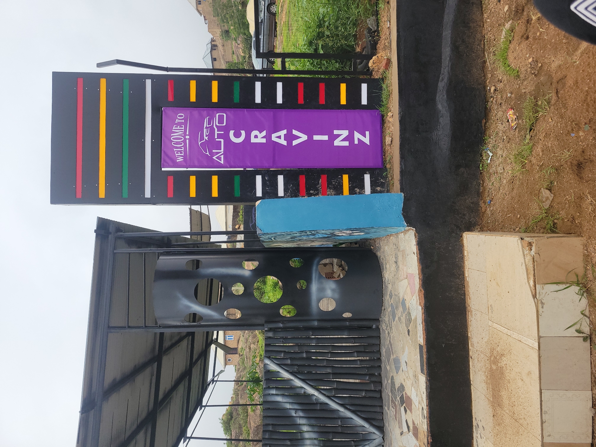 The image depicts an outdoor scene with a prominent sign and architectural elements. A large, dark gray rectangular structure stands to the right, featuring horizontal stripes of red, yellow, green, and white. In the center of this structure, a purple banner displays the text "WELCOME TO AUTO CRAVINN" along with a silhouette of a car. To the left of this sign is a black, cylindrical structure with circular cutouts, possibly a decorative pillar or part of a building. In front of the cylinder is a dark gray bamboo fence. A light blue, rectangular object, possibly a painted wall or a sculpture, is situated between the cylinder and the sign. The ground in front of these structures is paved with multi-colored tiles. In the background, a grassy area with some trees and buildings can be seen, suggesting a semi-urban or rural setting. The sky is overcast. The scene appears to be an entrance or a prominent feature of a commercial establishment.