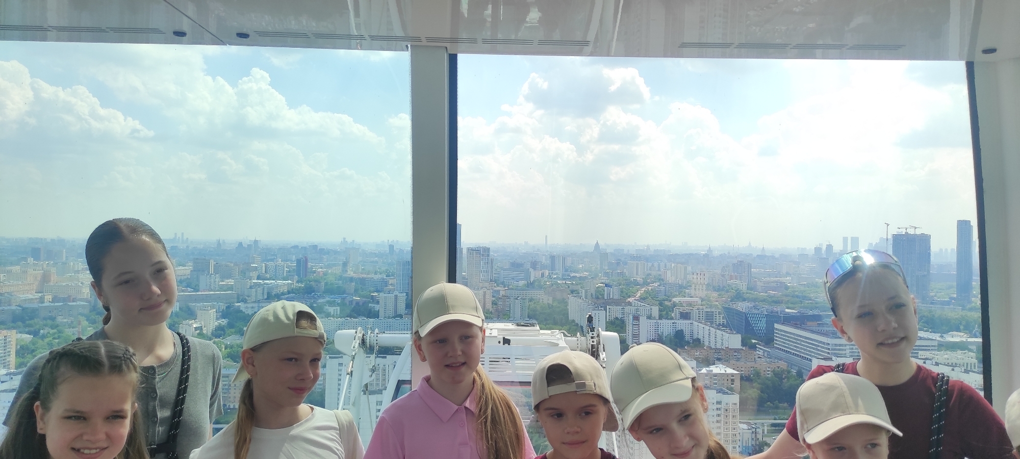 In the city of Khimki, Russia, eight young girls pose for a photo in a Ferris wheel gondola, overlooking a sprawling urban landscape under a partly cloudy sky. The girls, ranging in age from approximately six to fourteen years old, are positioned closely together. The girl on the far left, with braided hair, smiles directly at the camera. Next to her, a slightly older girl with dark hair tied back in a ponytail looks to her left, her expression neutral. Another girl in a pink polo shirt and a light-colored baseball cap is visible in the center, looking forward. To her right, a girl with a light-colored baseball cap and blonde hair pulled back smiles faintly. The girl on the far right, wearing mirrored sunglasses on her forehead and a dark red shirt, gazes towards the camera. Two other girls are partially visible in the foreground, one on the left and one in the center, with their faces obscured. The gondola's interior structure, including a white metal arm, is visible behind them, blending into the background. The city view is extensive, featuring numerous buildings of varying heights, trees, and distant skyscrapers, suggesting a high vantage point.