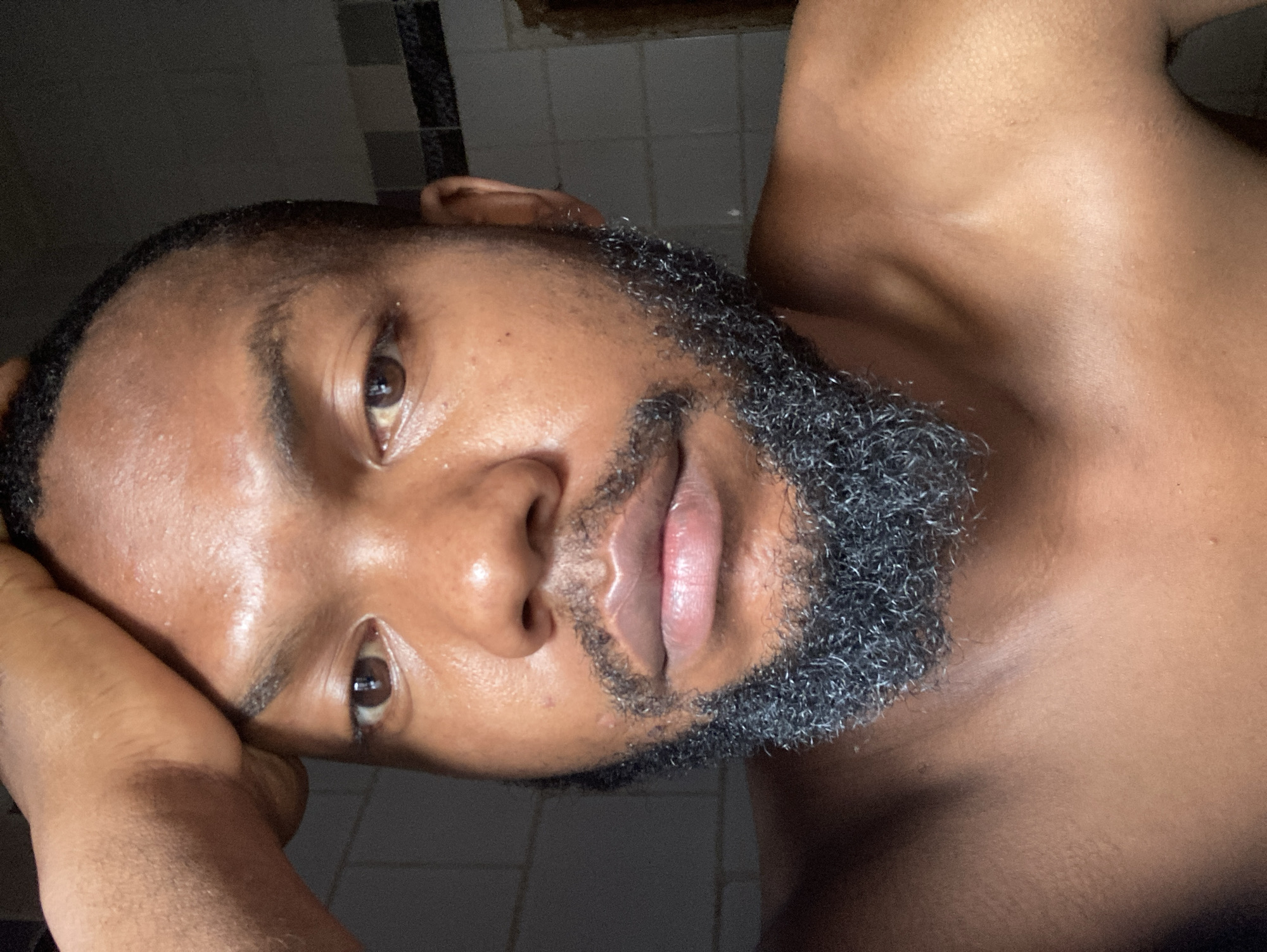 A close-up, low-angle portrait depicts a dark-skinned man with a graying, close-cropped beard. The man's skin appears shiny, and his lips are slightly parted. He is positioned at an angle, with his right arm seemingly supporting his head. The background shows out-of-focus elements of a tiled wall. There is no information available to place the subject in Jos, Nigeria.