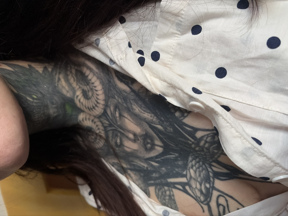 The image displays a close-up, downward-angled view of a person's upper torso, partially obscured by clothing and hair, revealing a large, intricate tattoo.
The person has dark, long hair that cascades over the scene, covering sections of the clothing and the tattoo. On the visible skin, a complex and detailed black and grey tattoo covers a significant portion of the chest or side. The tattoo features multiple interconnected elements. On the far left, a distinct, beast-like head with a sharp, angular profile is visible, distinguished by a bright, luminous green eye. Its scales or texture appear dark and rugged.
Adjacent to this beast's head and integrated into the overall design is what appears to be a stylized female face. This face has dark, possibly closed or downcast eyes, and is surrounded by flowing, intricate lines that resemble tendrils, snakes, or abstract hair, some of which form coiled or spiraling patterns. Further to the right and lower down in the image, more serpentine or scaled forms intertwine, suggesting a continuous and sprawling design. The tattoo's style is rich in detail and shading, creating a dark, fantastical, or gothic aesthetic.
The person is wearing a light-colored, possibly white or cream-colored, button-up shirt adorned with small, dark polka dots (likely black or navy blue). The shirt is slightly parted or unbuttoned, revealing the tattooed skin beneath. Several white buttons are visible along the placket of the shirt, one of which appears to be unfastened, contributing to the partial reveal of the tattoo. The fabric of the shirt shows some natural folds and creases.
The overall lighting appears soft and diffused, highlighting the details of both the tattoo and the fabric without harsh shadows. No specific activity is taking place; the image is a static shot focused on presenting the body art. There is no visible text in the image.