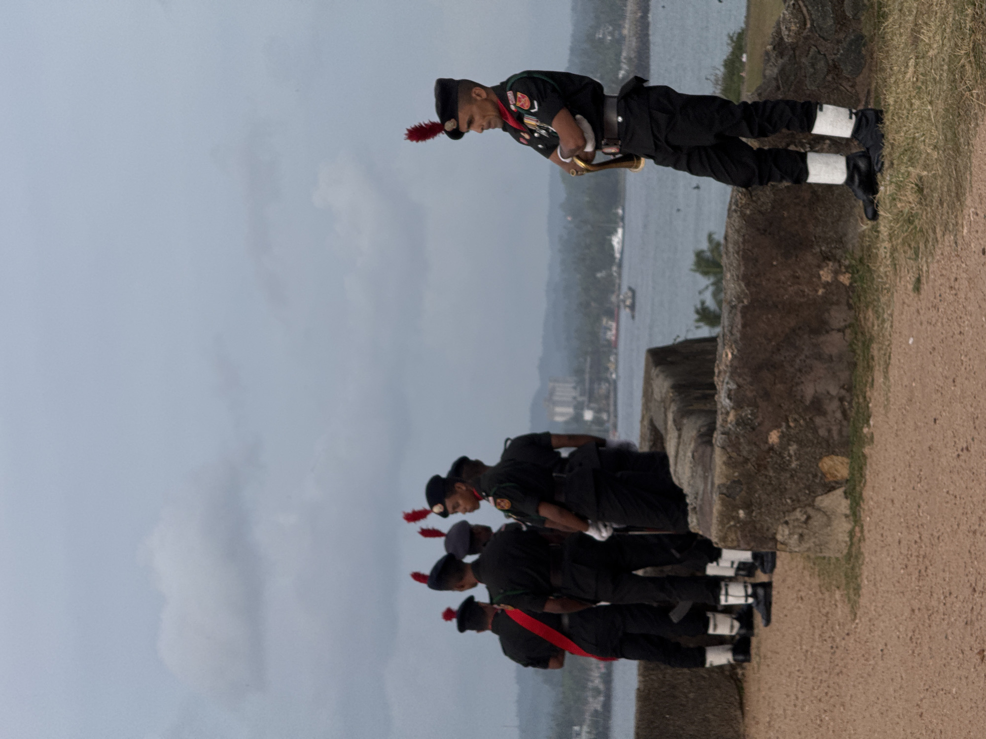 A group of men in military uniform stand guard at Galle Fort, Sri Lanka, a UNESCO World Heritage Site. The men, all wearing black uniforms with red sashes, red plumes on their hats, and white spats on their boots, are positioned near a low stone wall. One man in the foreground stands with his arms crossed, facing away from the viewer. The background shows a body of water, likely the Indian Ocean, with boats in the distance and a hazy cityscape. The sky is overcast, suggesting a cloudy day. The overall scene depicts a military presence within the historic fortifications of Galle.