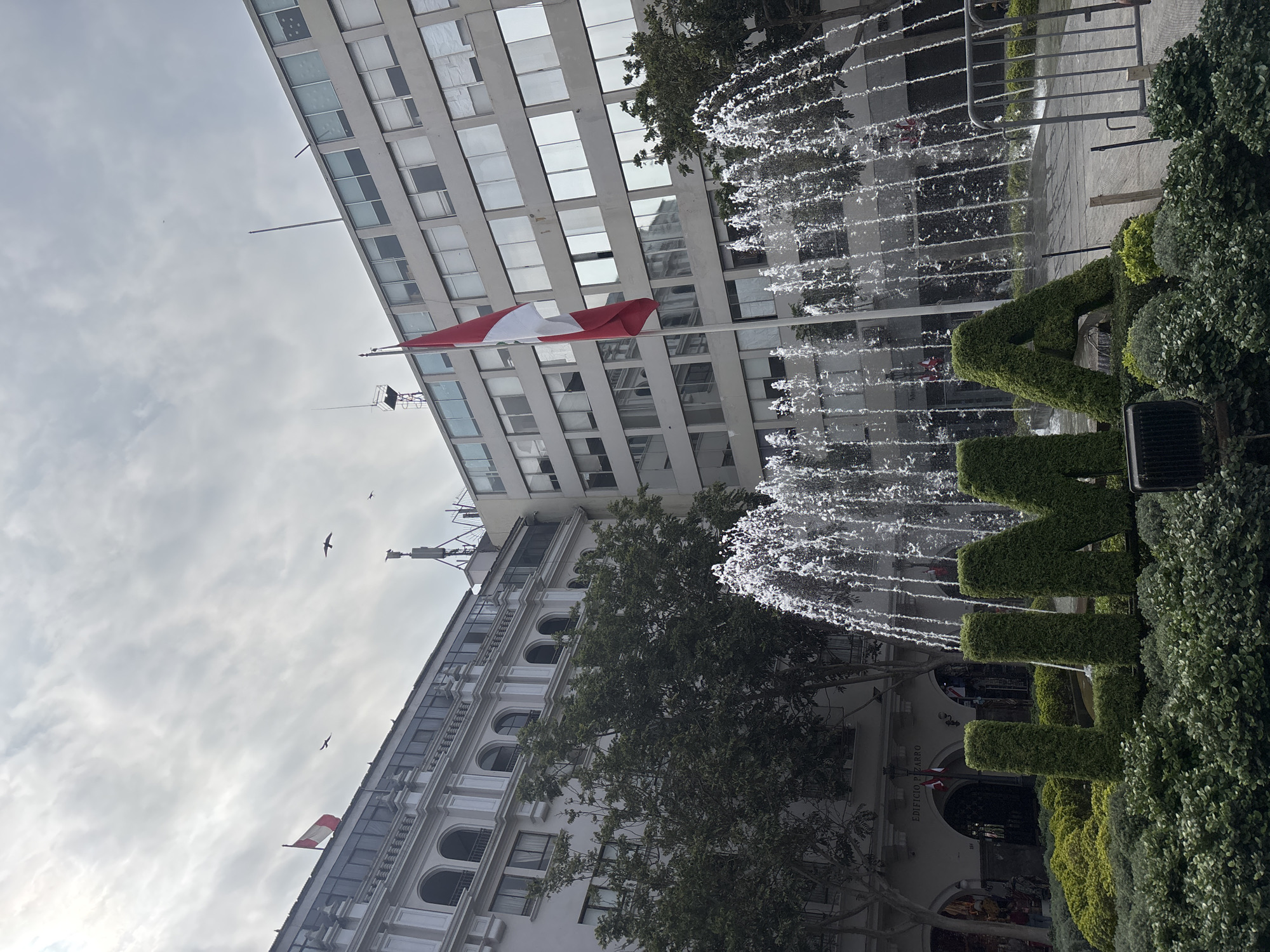 This image depicts the Plaza de Armas in Lima, Peru, featuring a prominent fountain with water cascading down. In the foreground, sculpted hedges form the letters "LIMA". Behind the fountain, a large, modern building with many windows dominates the skyline. To the left, an older, ornate building with arched windows is visible. A Peruvian flag flies from a flagpole in front of the modern building, and another smaller flag is seen on the older building. Several birds are in flight against a cloudy sky. Trees with green foliage are interspersed between the buildings. The scene appears to be a public square in an urban setting.