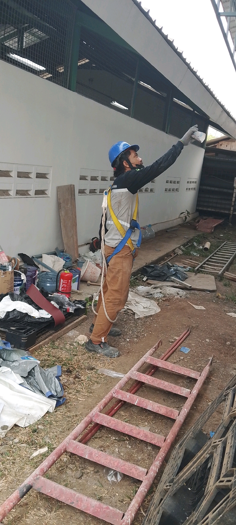 A male worker, wearing a blue hard hat, a yellow safety harness, and brown work pants, is standing outdoors and pointing at a building. He is holding a white object in his gloved right hand, possibly a spray can or a document. The background shows a white building with vents and a metal roof structure. Various construction materials and debris, including a red ladder, buckets, and tarps, are scattered on the ground. The scene appears to be in an industrial or construction area, likely within the city of Depok, Indonesia, given the context. The lighting suggests it is daytime, possibly overcast.