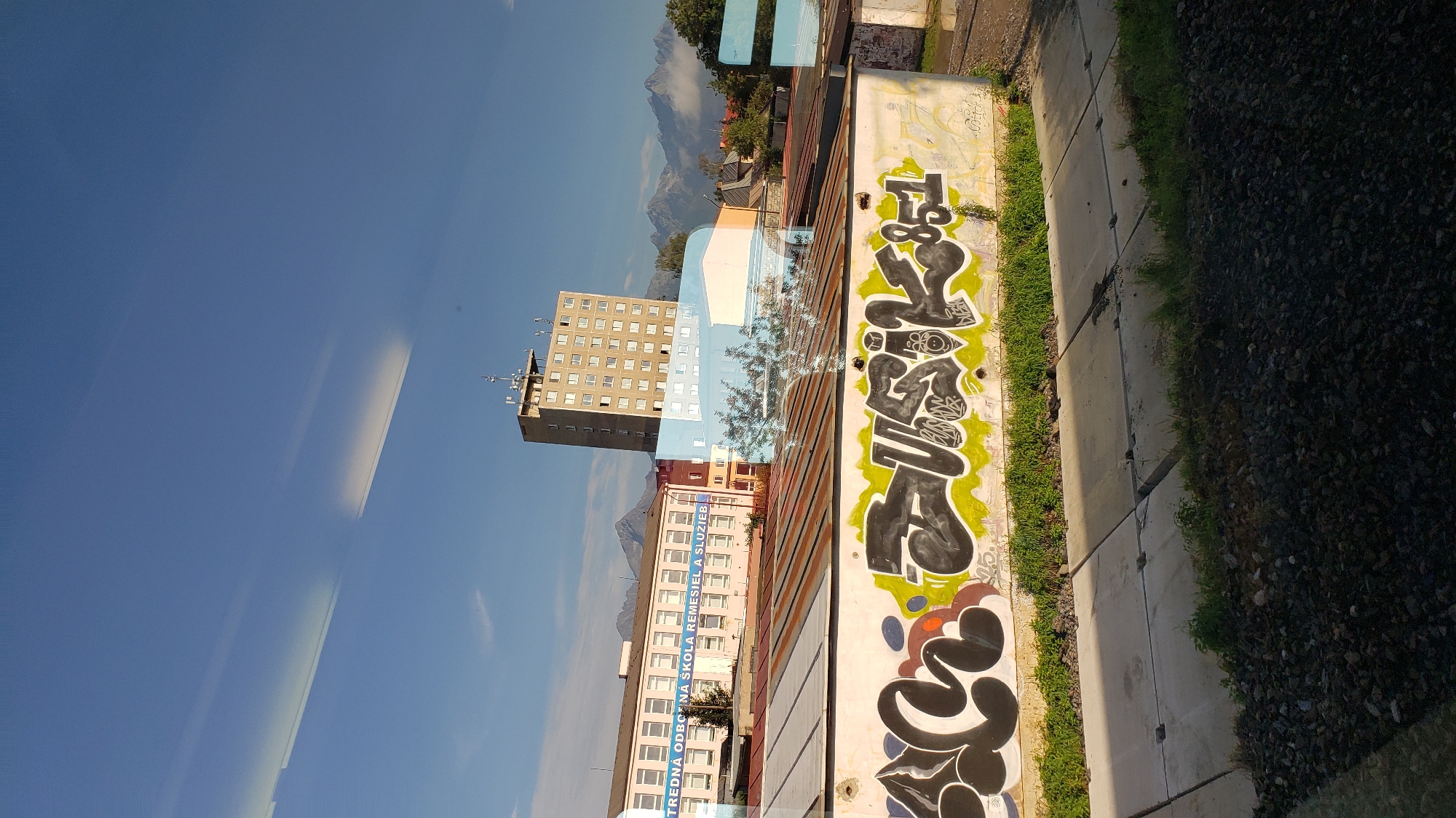 The image is a photograph taken from a moving vehicle, likely a train, in Poprad, Slovakia. The foreground shows a gravel track bed and concrete slabs, suggesting the perspective is from a train window. In the midground, a white wall is covered with graffiti, featuring black and lime green lettering. Beyond the wall, there is a flat, corrugated metal roof and a light pink building with a banner that reads "STREDNÁ ODBORNÁ ŠKOLA REMESIEL A SLUŽIEB". To the right of this building is a tall, grey, multi-story structure with many windows and an antenna on top. The background features a clear blue sky with some streaky clouds and distant, snow-capped mountains, characteristic of the Tatra Mountains region. The scene depicts an urban or industrial area with a backdrop of natural mountain scenery.