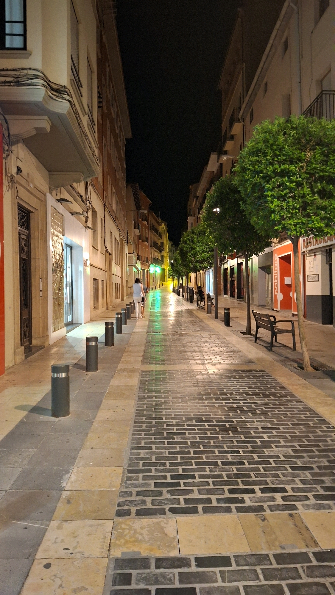 This is an outdoor, eye-level, full-frame shot of a pedestrian street in Alcañiz, Spain at night. The street is paved with a combination of large tan and grey stone slabs and darker brickwork in a herringbone pattern. The street is lined with buildings on both sides, which are illuminated by artificial lights.
On the left side of the street, there are storefronts with large glass windows, one of which displays a brightly lit interior. Several dark cylindrical bollards are spaced along the edge of the sidewalk.
On the right side, there are more buildings with various colored facades. Three ornamental trees with full green foliage are planted along the sidewalk. There are two dark wooden benches positioned along this side of the street. One bench is occupied by two individuals. Further down the street, another bench appears to be occupied by a single person.
A person wearing a light blue top and white pants is walking away from the camera down the center of the street, followed by another person. The street is wet, reflecting the green and yellow lights from overhead lamps, creating a luminous effect on the pavement. In the distance, the street appears to curve and continue between buildings under a dark night sky. A sign that reads "RESTAURANT" is visible on one of the buildings on the right.