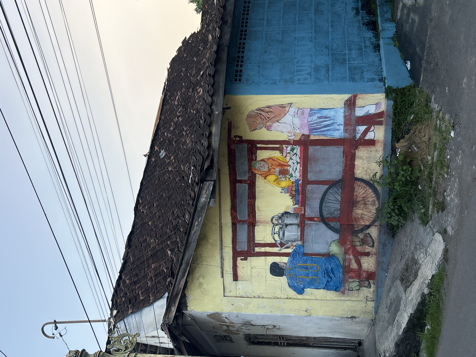 This image shows a mural painted on the side of a building. The mural depicts a scene with three people. One person, wearing a pink hijab and blue pants, stands behind a stall. Another person, wearing an orange hijab and yellow shirt, is seated behind the stall. A third person, wearing a blue jacket, is seated in front of the stall, facing away from the viewer.
The stall itself is made of red wood and has a bicycle wheel attached to one side. There are several metal containers on the stall. The building has a yellow lower section and a blue upper section. A dark brown tiled roof extends over the mural.
The scene appears to be set in a street or alleyway. Overhead, a tangle of electrical wires is visible against a pale sky, suggesting it is daytime, possibly late afternoon or early morning given the soft light. The ground in front of the building is paved with concrete and asphalt, with some patches of grass and dry vegetation. The overall impression is one of a bustling street scene, possibly depicting vendors at a market or food stall. The mural has a slightly weathered appearance, with some paint peeling on the lower part of the building. There is no visible text in the image.
