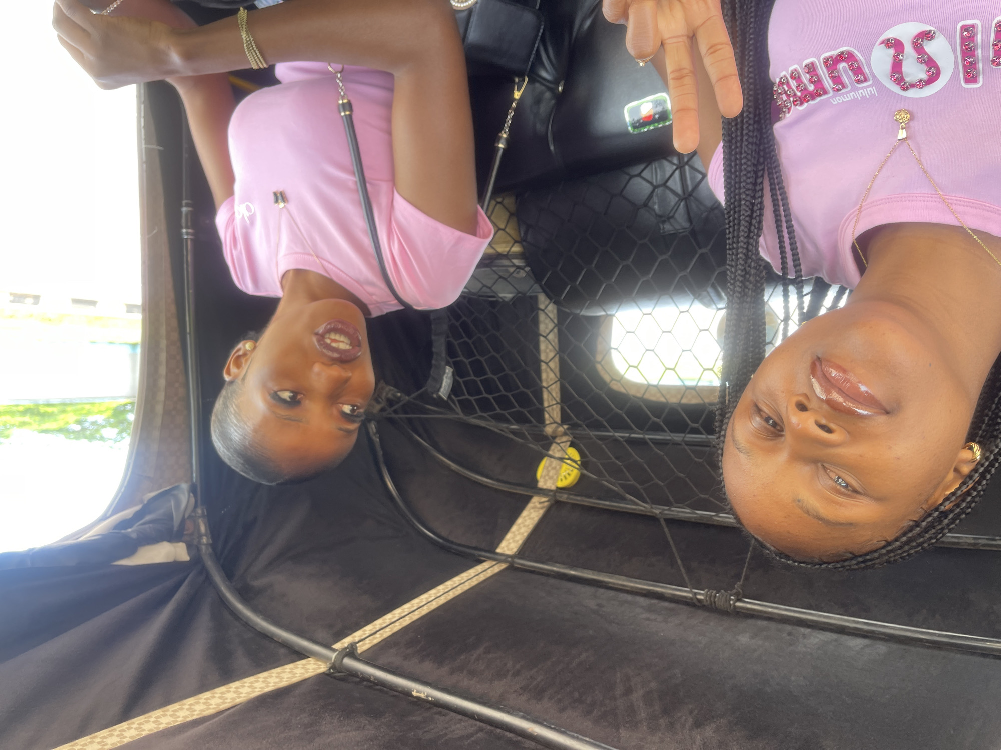 Two young women are inside a vehicle, possibly an auto-rickshaw or tuk-tuk, which is common in Barakin Lerawi, Nigeria. The woman on the left, closer to the camera, has dark braided hair and is wearing a pink t-shirt with a logo and a gold necklace. She is looking at the camera and holding up two fingers in a peace sign. The woman on the right has her hair pulled back and is wearing a similar pink t-shirt and a gold necklace. She is looking to her right and her mouth is open, as if she is speaking. Both women appear to be of African descent. The interior of the vehicle is dark with some light filtering in from the windows. The background outside the vehicle is bright and appears to be a street scene with some greenery. The overall impression is a candid, casual moment captured between two friends.
