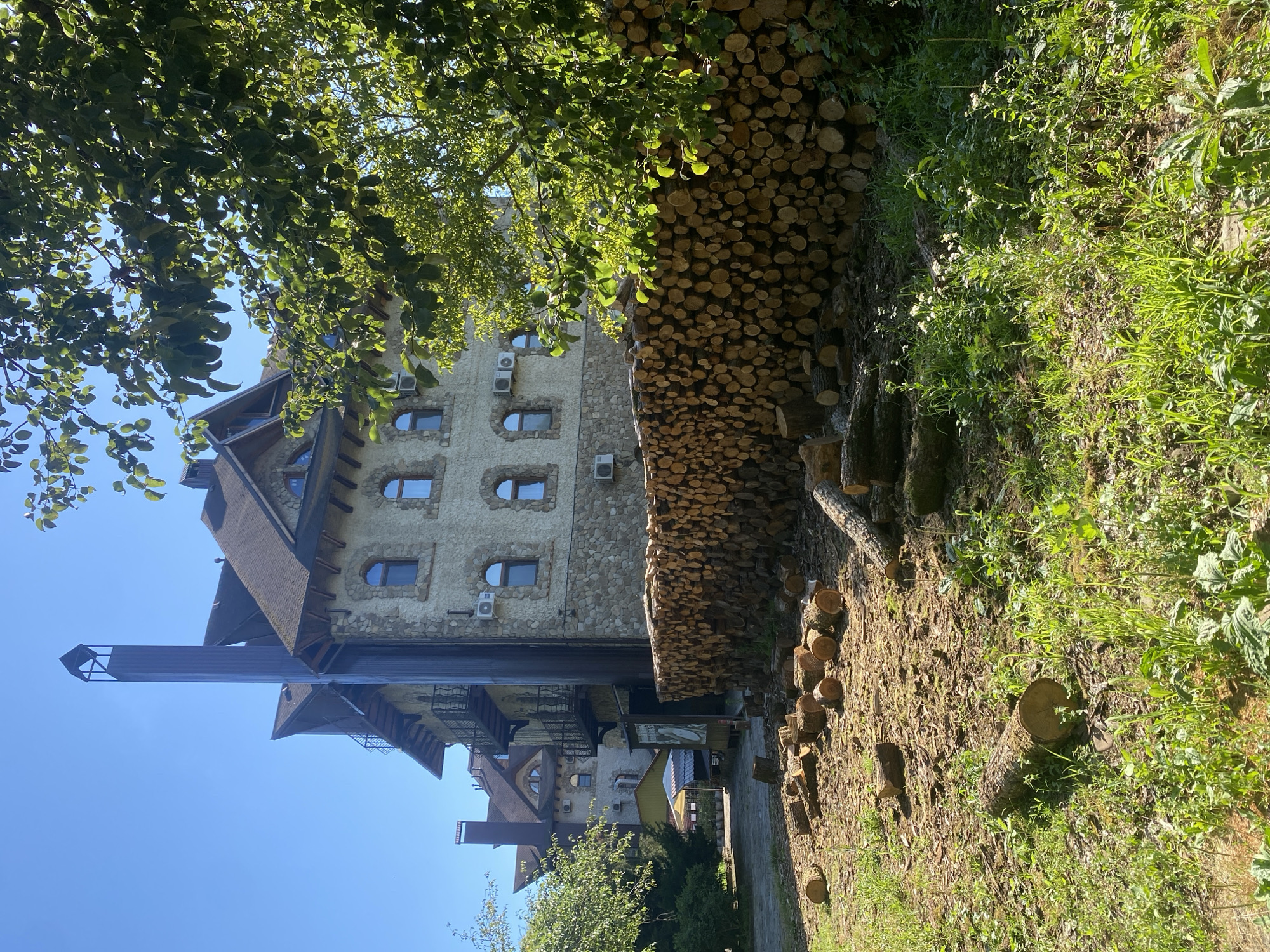 The image depicts a building with a stone facade and multiple windows, set against a clear blue sky. In front of the building, a large pile of cut logs is stacked. Scattered on the ground in front of the log pile are additional logs and branches. Trees with green foliage are visible on the right side of the image, partially obscuring the view of the building. In the background, there are other buildings and structures, including what appears to be a covered seating area with a yellow roof. The overall scene suggests an outdoor environment, possibly a residential or commercial property with a focus on wood storage. The location is identified as Goryachiy Klyuch, Russia. There are no people or animals visible in the image.