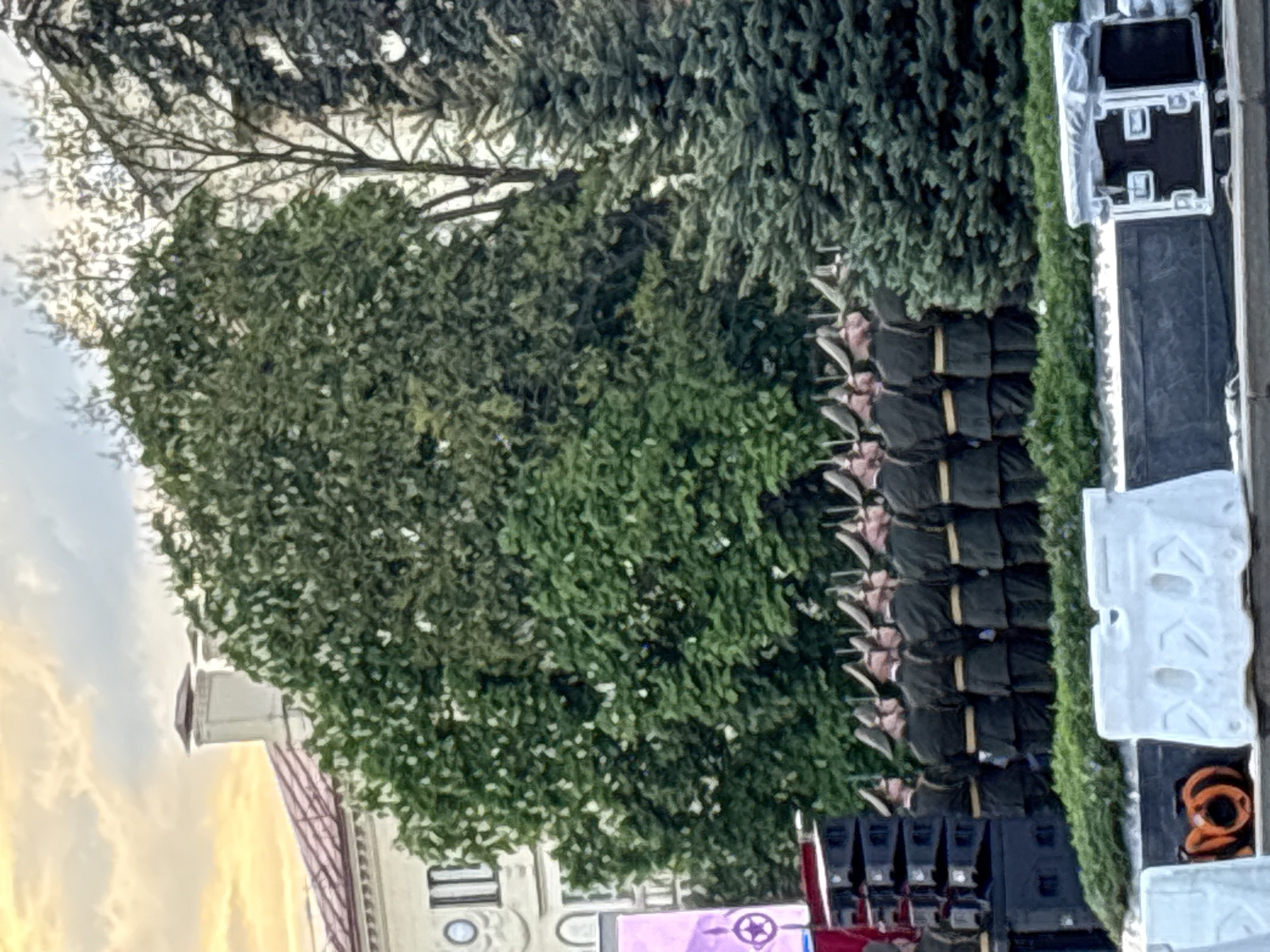 The image depicts a military parade formation in Minsk, Belarus. The soldiers are standing in formation and saluting. Adjacent to the parade formation is an area with trees, event support equipment, and barriers. In the background, there is a building with a tower, partially obscured by trees. The sky is visible with light clouds and suggests daylight.