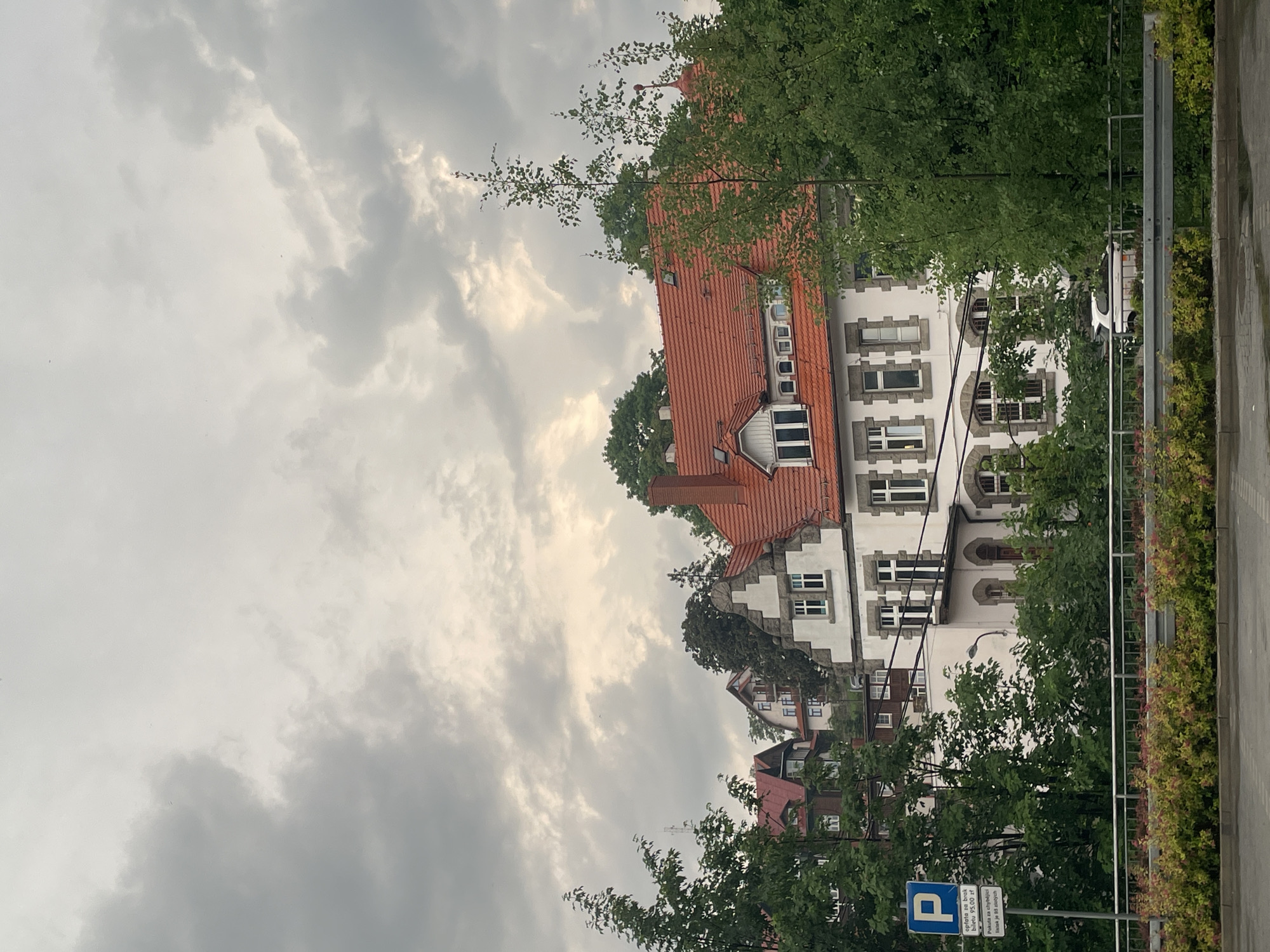 The image shows a view of buildings and greenery under a cloudy sky in Szklarska Poręba, Poland. In the foreground, a gray metal guardrail runs horizontally across the frame, with lush green and reddish-yellow bushes planted behind it. Beyond the guardrail, a white car is partially visible.
Dominating the middle ground is a multi-story building with a prominent red-tiled roof. The building has white walls with stone accents around the windows and entrances. Several windows are visible, some rectangular and others arched. Balconies with railings are also present. To the left of this main building, other buildings with red roofs and similar architectural styles are visible on a hillside, some appearing older and partially obscured by trees.
The right side of the image is filled with dense green foliage and trees, some of which obscure parts of the buildings. On the left side of the main building, a blue and white parking sign is mounted on a pole. The sign features a white "P" on a blue background, with text below it, likely related to parking regulations or payment.
The sky above is heavily overcast with dramatic gray clouds, with patches of lighter sky suggesting diffused sunlight. The overall impression is of a slightly somber or overcast day in a residential or institutional area surrounded by nature.