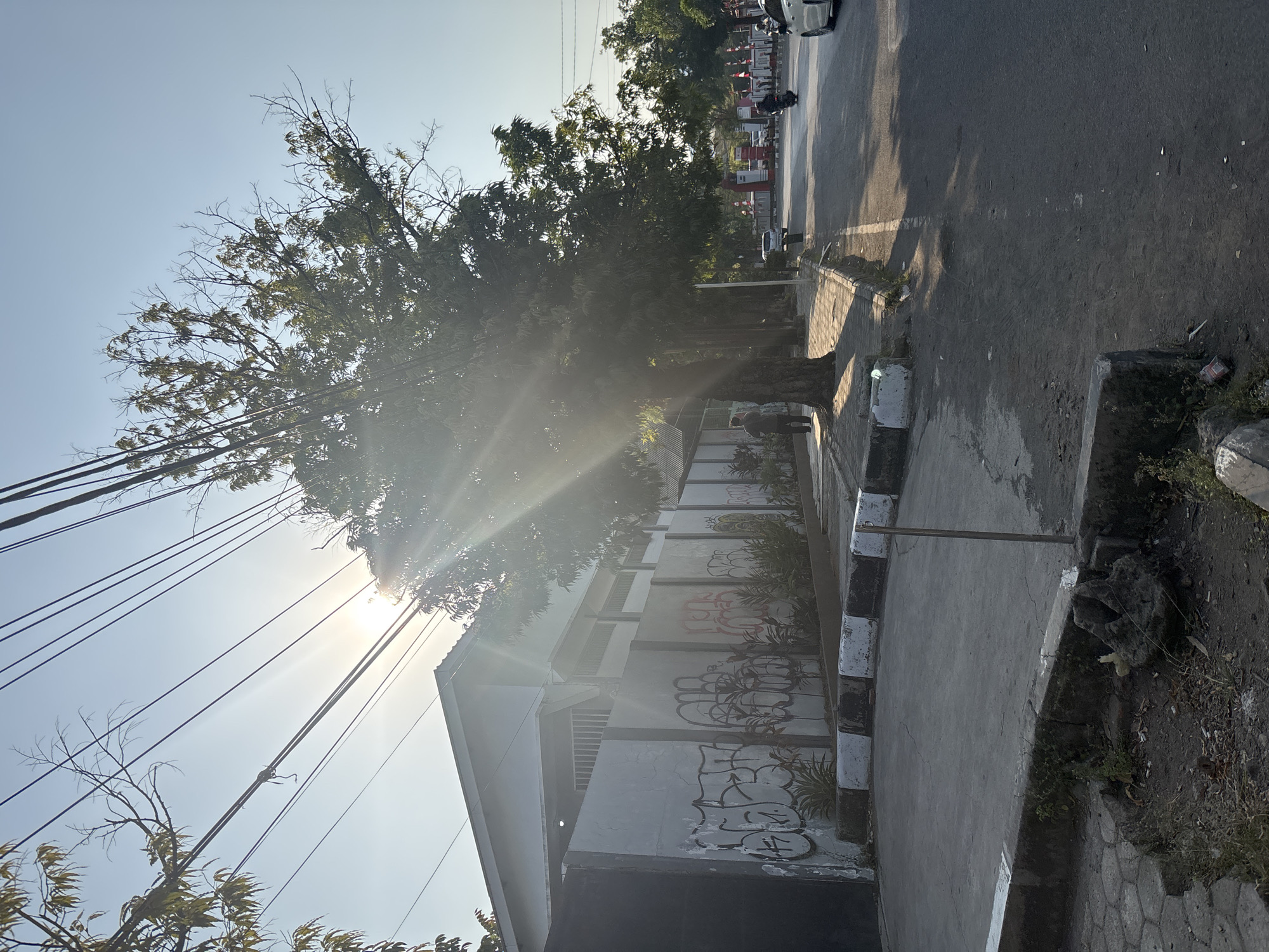 The image captures a sun-drenched street scene in Cirebon, Indonesia, likely in a commercial or residential area with a mix of buildings and vegetation. The sun's rays are prominently featured, creating lens flare and casting long shadows across the pavement and sidewalk.
On the left side of the frame, a long, gray concrete wall is visible, adorned with graffiti. Behind the wall, the roofline of a building is discernible. A large tree with dense foliage stands near the sidewalk, its branches extending over the street. Power lines crisscross the sky, further emphasizing the urban setting.
The sidewalk in front of the wall is paved and has a border marked with black and white stripes. A person is walking on the sidewalk near the large tree, appearing as a dark silhouette against the bright sunlight.
The street itself is asphalt, with a white line marking the edge of the sidewalk. Several vehicles are present on the street, including a motorcycle with a rider and a white car. In the distance, there are signs and structures that suggest commercial activity, possibly a gas station or a row of shops, indicated by red and white signage.
The overall impression is that of a typical day in a bustling Indonesian city, with the bright sunlight creating a dramatic effect on the ordinary urban landscape. The presence of graffiti and power lines indicates a lived-in, functional environment.