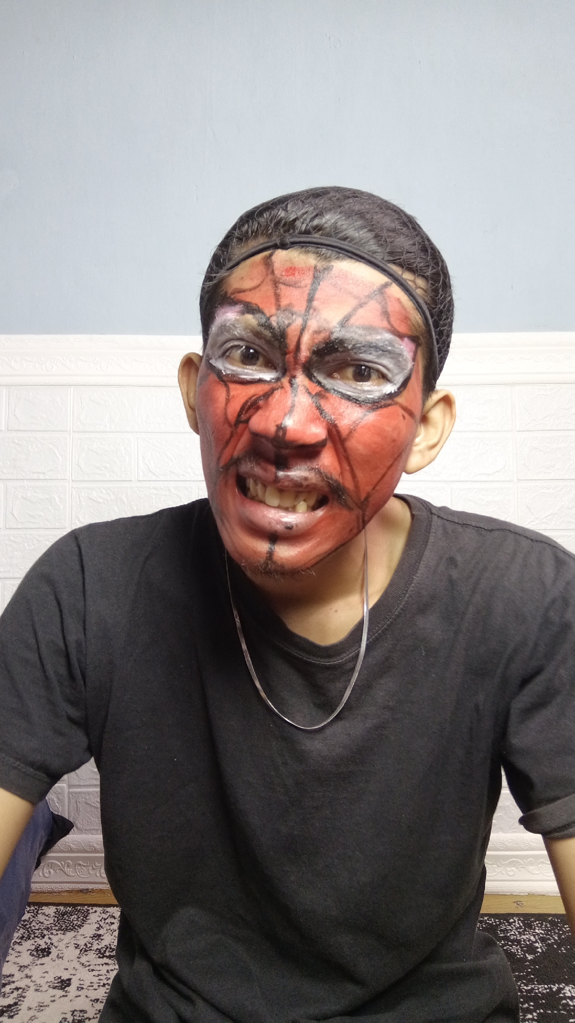 A person with Spider-Man-themed face paint is looking at the camera. The paint is red with black lines and white around the eyes. The person is wearing a black t-shirt and a silver chain necklace. The background consists of a white textured wall and a light blue wall. The person is sitting, as evidenced by the visible rug and furniture. The image was taken indoors in Kebumen, Indonesia.
