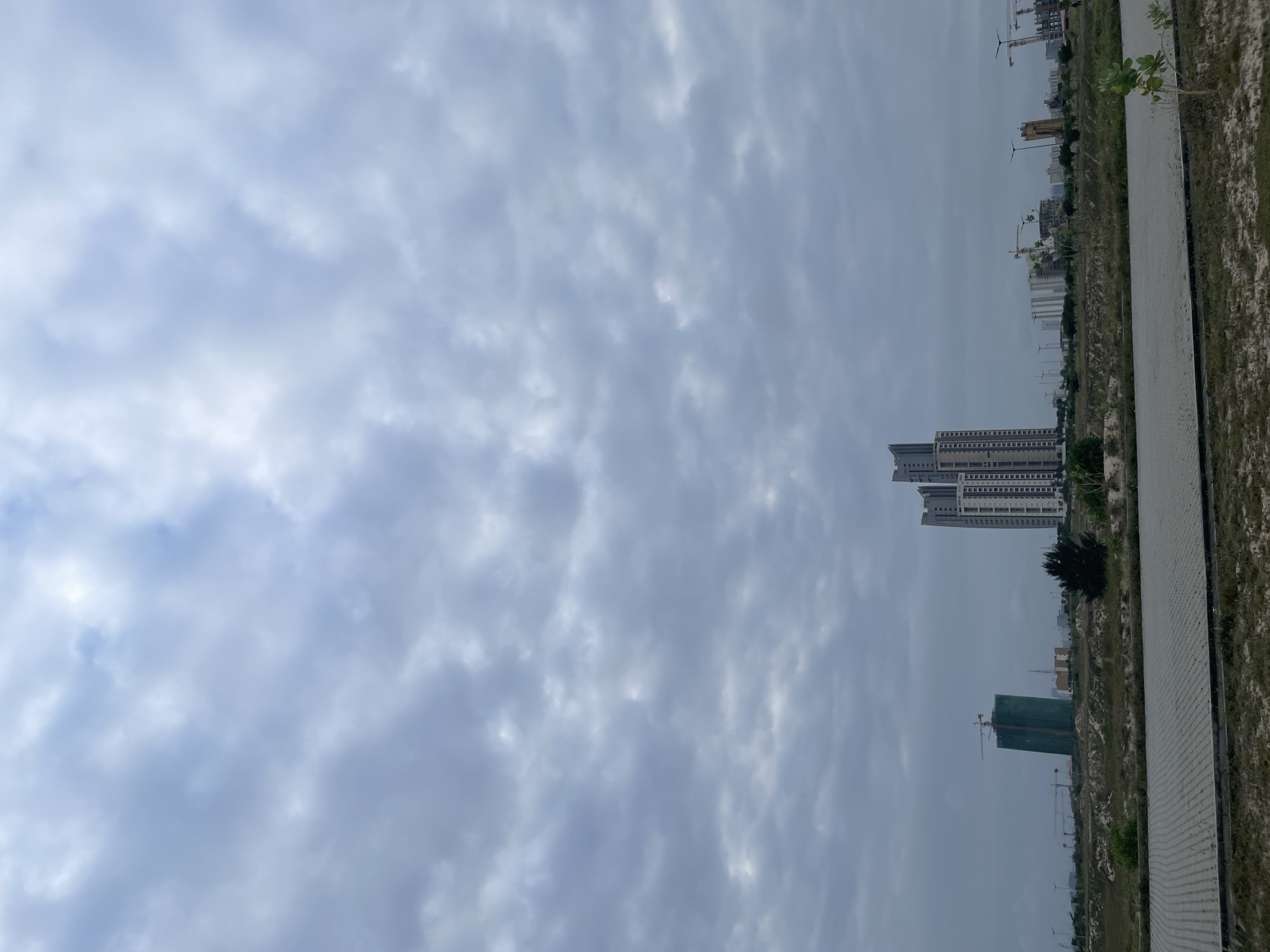 The image depicts an overcast sky above a developing urban area, likely in Lagos, Nigeria. In the foreground, a paved walkway is bordered by dry grass and sparse vegetation. In the mid-ground, the landscape transitions to undeveloped land with patches of sand and scrub. A prominent feature is a tall, modern skyscraper with multiple sections and a grey facade, standing to the right of center. To its left, a partially constructed building is covered in green netting, with a crane visible above it. Further in the background, a line of other buildings, including more high-rises and construction cranes, suggests ongoing development. The overall scene conveys a sense of urban expansion under a cloudy sky.