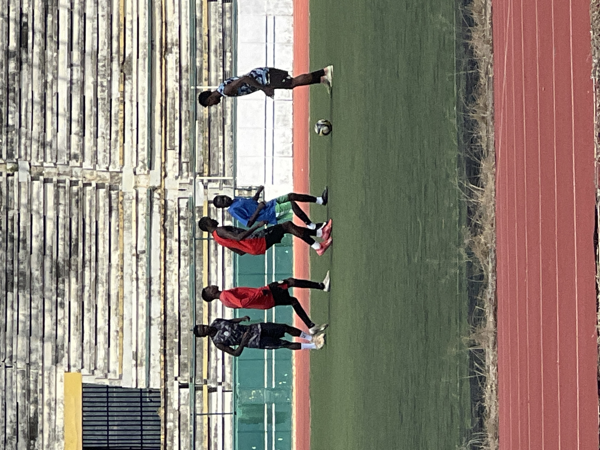 At the New Stadium in Nigeria, five young men are engaged in a soccer practice session on a green turf field. Two men, wearing red jerseys and black shorts, are in the center of the frame. To the left, a man in a grey and black patterned jersey and black shorts stands looking on. To the right, a man in a blue jersey and green shorts is positioned with his back to the camera. Further to the right, another man in a blue, black, and white patterned jersey and black shorts is facing the group. A soccer ball rests on the grass between the two men on the right. In the background, a multi-tiered grandstand with concrete seating and dark metal railings is visible, along with a dark metal fence and a green barrier along the edge of the field. A red running track borders the lower part of the image, with dry grass and vegetation separating it from the soccer field.