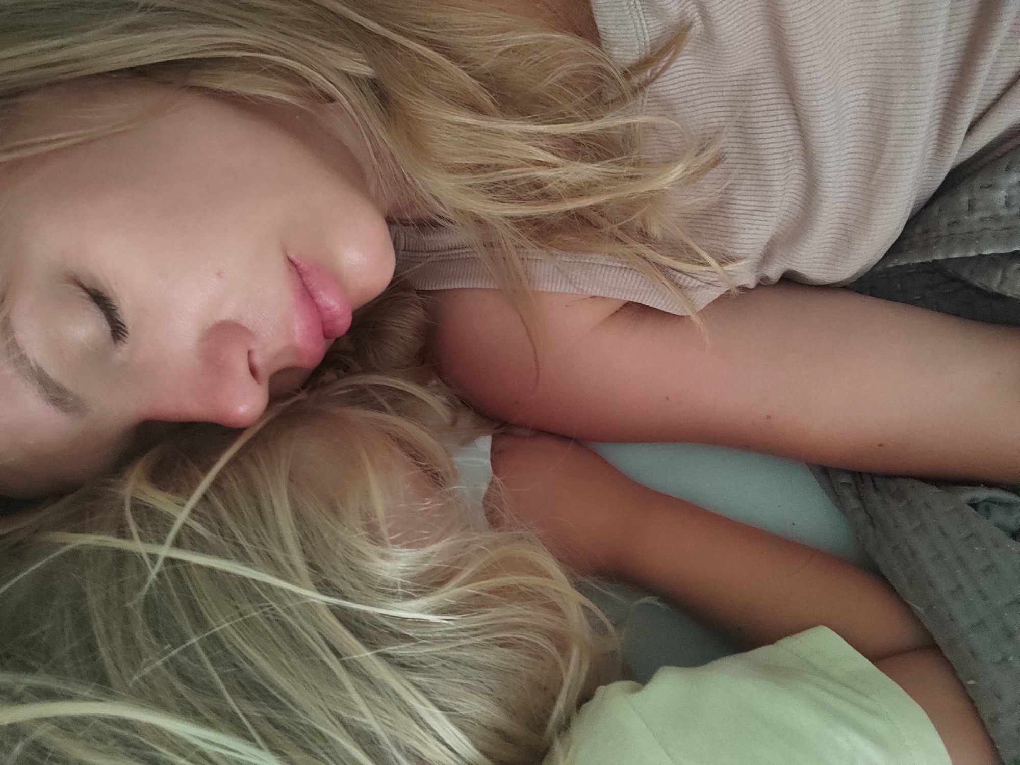 The image shows a close-up, overhead view of two people sleeping. The person in the foreground, whose face is partially visible, has blonde hair and is wearing a light pink, ribbed top. Their eyes are closed, and their lips are slightly parted. Behind them, another person with blonde hair is visible, also sleeping. This person is wearing a light yellow t-shirt. They are lying on a grey, textured blanket. The overall scene suggests a moment of rest or sleep. The image does not contain any elements that indicate its location in Blanes, Spain.