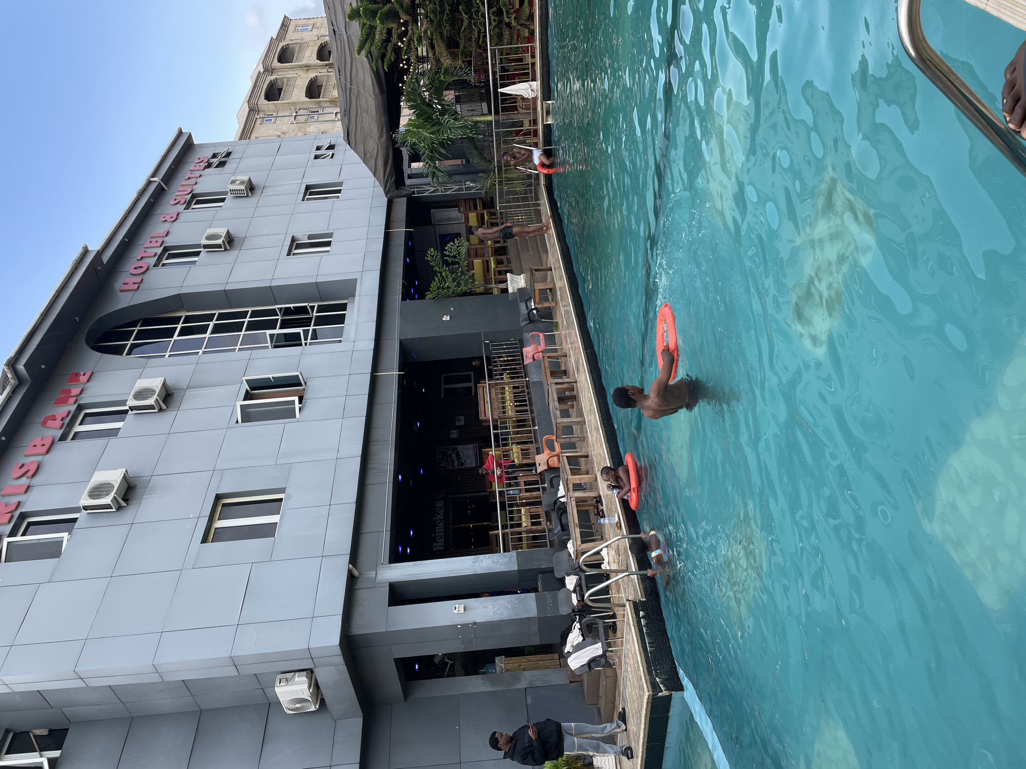 The image shows a poolside scene at a hotel in Nkpor, Nigeria. The hotel building, with its grey facade and red lettering spelling out "HOTEL & SUITES," is visible on the left. Several air conditioning units are mounted on the exterior walls, and windows punctuate the building's structure.
In the foreground, a bright turquoise swimming pool dominates the right side of the frame. Several people are in the pool, with one person in the water holding an orange flotation ring. Another person is standing near the edge of the pool, looking into the water. The poolside area features metal railings and wooden furniture, suggesting it's a leisure area. In the background, more of the hotel building and some greenery are visible, along with what appears to be an outdoor seating area with tables and chairs. The lighting suggests it is daytime.
The overall mood appears relaxed and casual, typical of a hotel environment with a swimming pool. No specific emotions are strongly conveyed, but the presence of people enjoying the water indicates leisure activity. The weather appears clear and sunny.