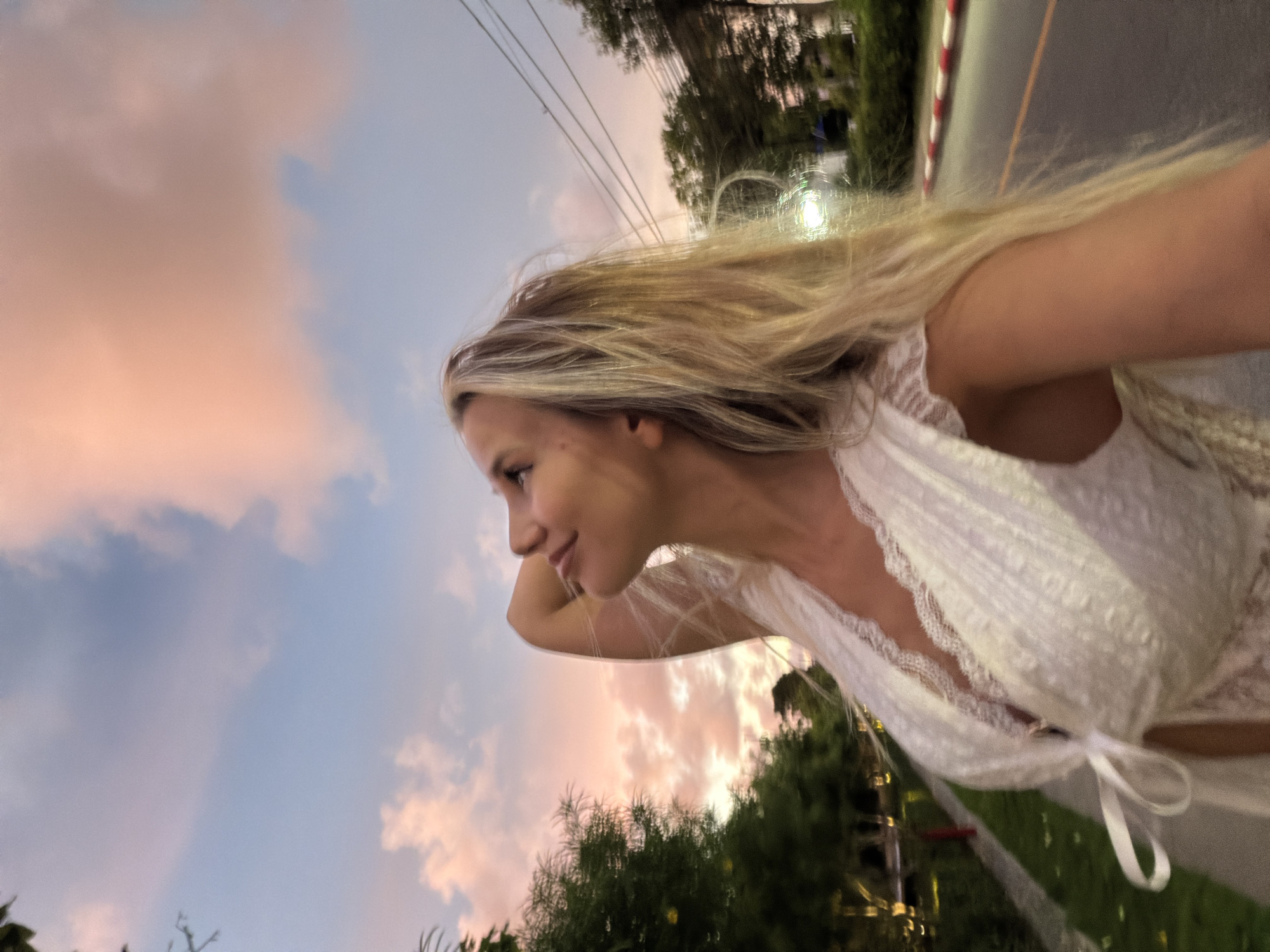 A blonde woman in a white lace dress is looking up at a colorful sunset sky. She is standing on a sidewalk next to a street with a red and white barrier. Trees and power lines are visible in the background. The scene appears to be in a tropical or sub-tropical location. The woman's right arm is raised behind her head. The street is illuminated by streetlights. The sky has hues of pink, orange, and blue with scattered clouds.