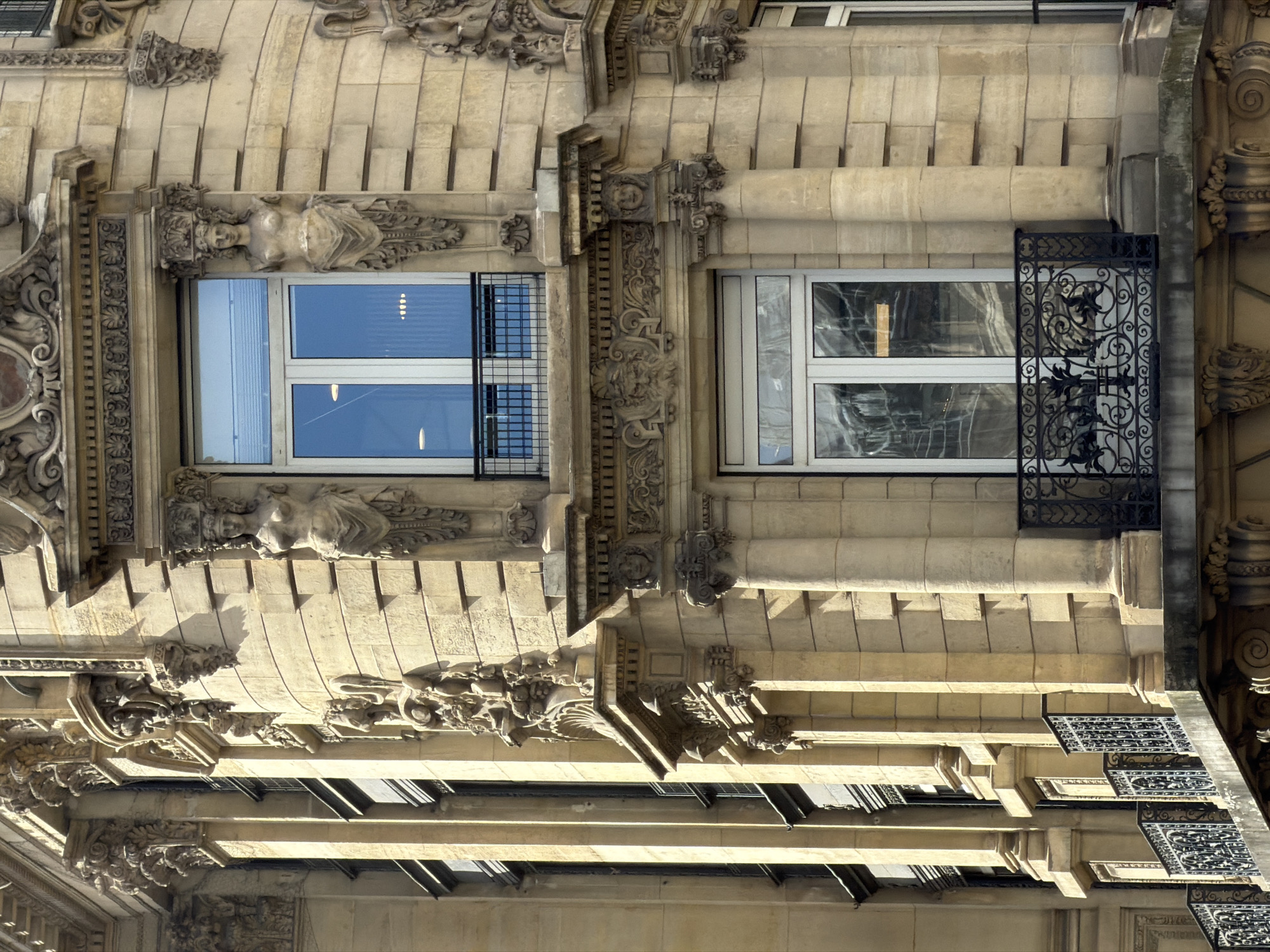 The image shows a detail of a Parisian building facade, likely in the Haussmann style. The facade is made of beige stone and is adorned with intricate architectural details such as cornices, moldings, and sculptures. There are two windows visible, both with white frames. The window on the left has a metal security grille in front of it, while the window on the right has a decorative ironwork screen. The building appears to be well-maintained and is likely located in a historic district of Paris.