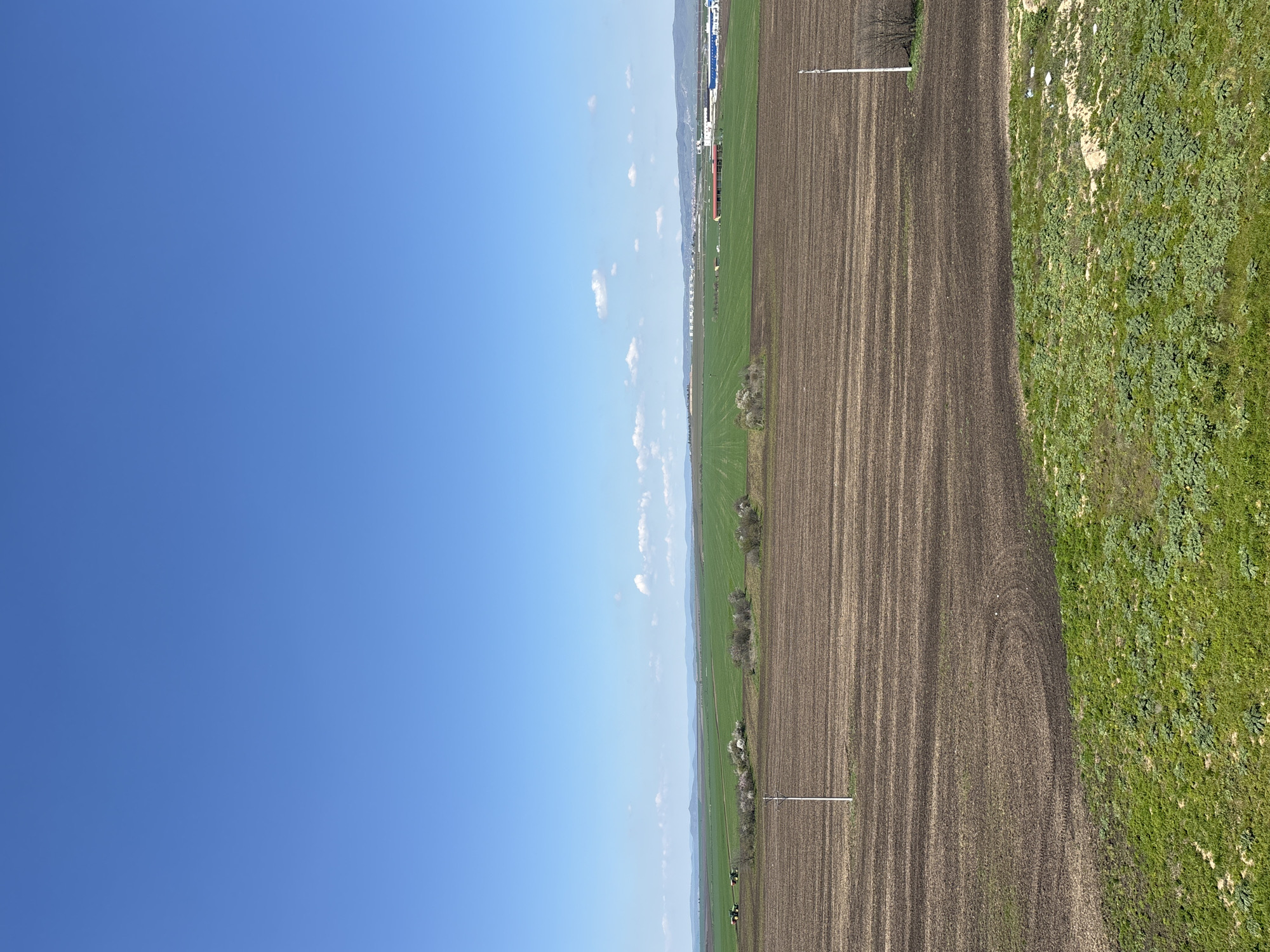 The image shows a rural landscape, possibly located in the Burgas area, Bulgaria. The left side of the image is dominated by a clear blue sky with scattered clouds. The right side features agricultural fields: one brown field with visible plow lines, another field that is green, and a strip of uncultivated land covered with green vegetation. Two white poles are situated at approximately equal distances from the bottom edge of the image. Mountains or hills are faintly visible in the distance along the horizon. Two tractors are present on the green field.