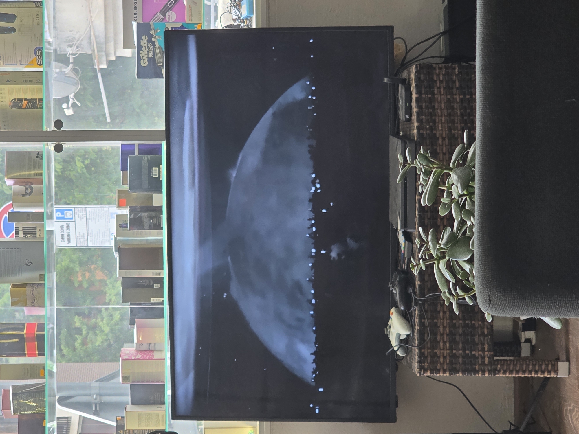 The image depicts a scene in what appears to be a retail store. In the foreground, a large television screen displays a dark, cloudy image with scattered lights, possibly a video game or movie scene. Below the TV, a dark wicker stand holds two video game controllers, one white and one black, along with some other electronic devices and cables. A potted plant with thick, fleshy leaves is positioned in front of the stand and a dark grey sofa.
In the background, shelves filled with various boxes, likely perfume or cosmetic products, are visible. Some of these boxes are brightly colored, while others are more subdued. Through a window or glass partition behind the shelves, greenery and trees can be seen, suggesting an outdoor environment. A sign with Cyrillic lettering, including "СИНЯ ЗОНА" (Blue Zone) and "PARKING ZONE," is also visible, indicating that the location is likely in a city with paid parking, such as Varna, Bulgaria. The overall impression is a retail space that also serves as a personal entertainment area.