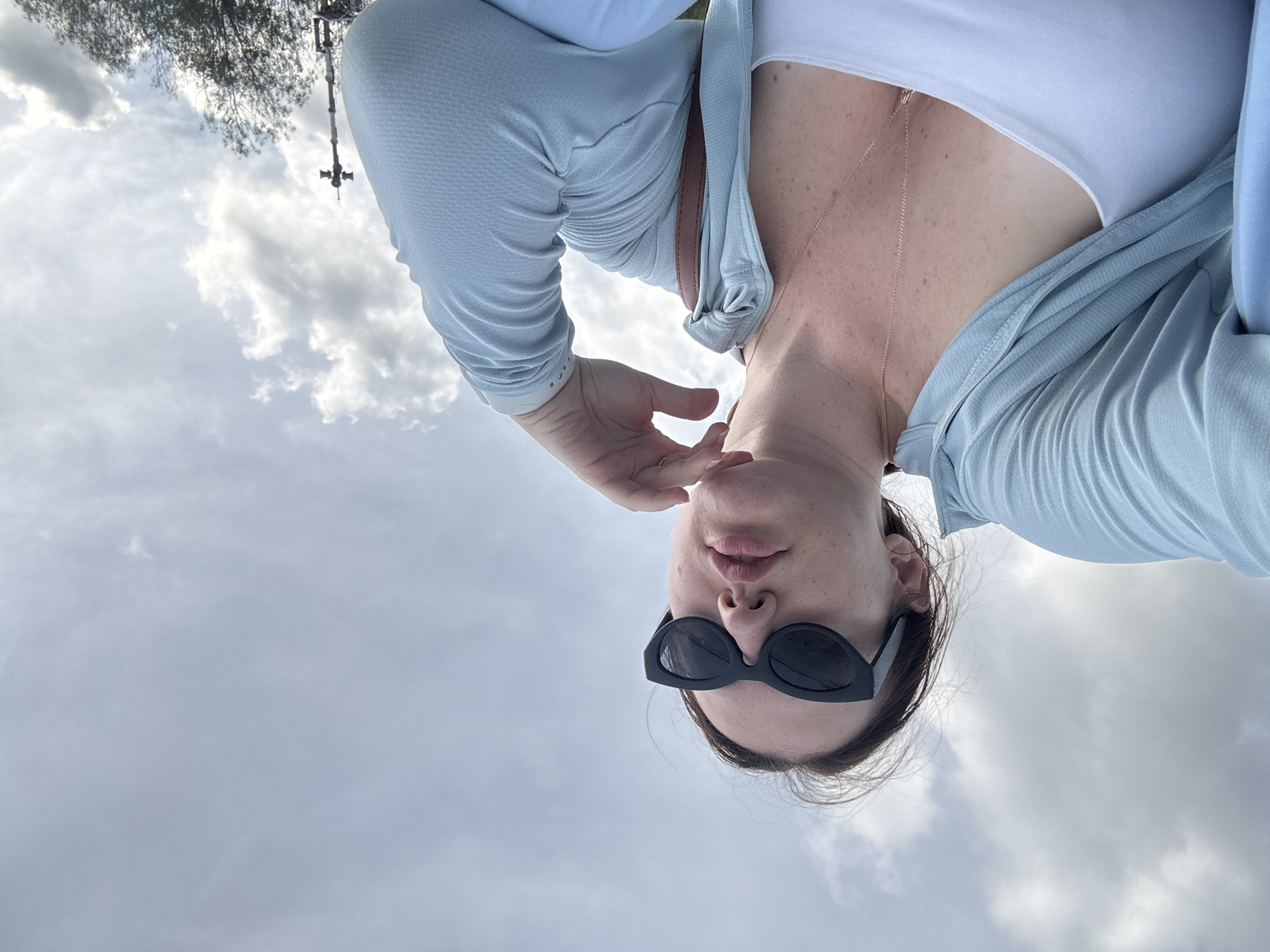 An extreme low-angle, outdoor, selfie-style photograph captures a woman sitting outside, looking up and to the right. She is wearing a light blue hooded zip-up jacket over a white tank top and black, cat-eye sunglasses. Her right hand is resting on her chin, and she has a single gold necklace. The background is an overcast sky with scattered clouds. To the far right of the image, a tall metal pole with two speakers on top is visible, along with the tops of trees. The photograph was taken in Phuket City Municipality, Thailand.