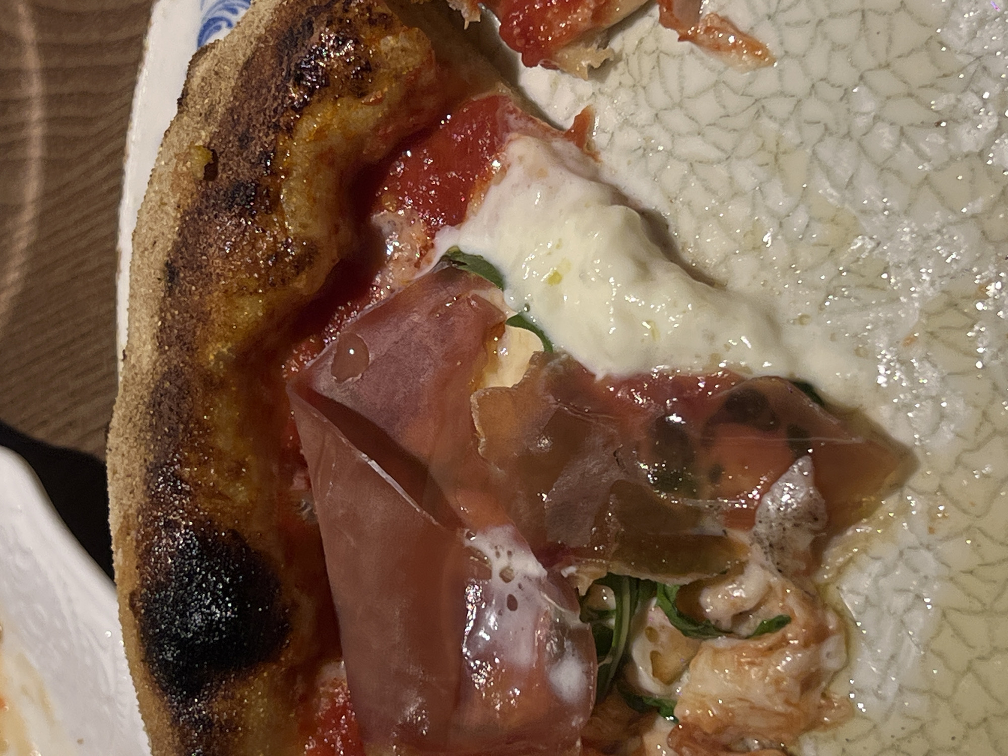 A close-up shot depicts a partial slice of pizza on a white plate with a patterned design. The pizza features a thick crust with a burnt appearance on the edge. Toppings include red tomato sauce, melted white cheese, prosciutto, and green arugula. The location and city are unknown. The main subject is the slice of pizza. There are no people visible in the frame. The action is the presentation of the pizza. The environment appears to be indoors, likely a restaurant setting.